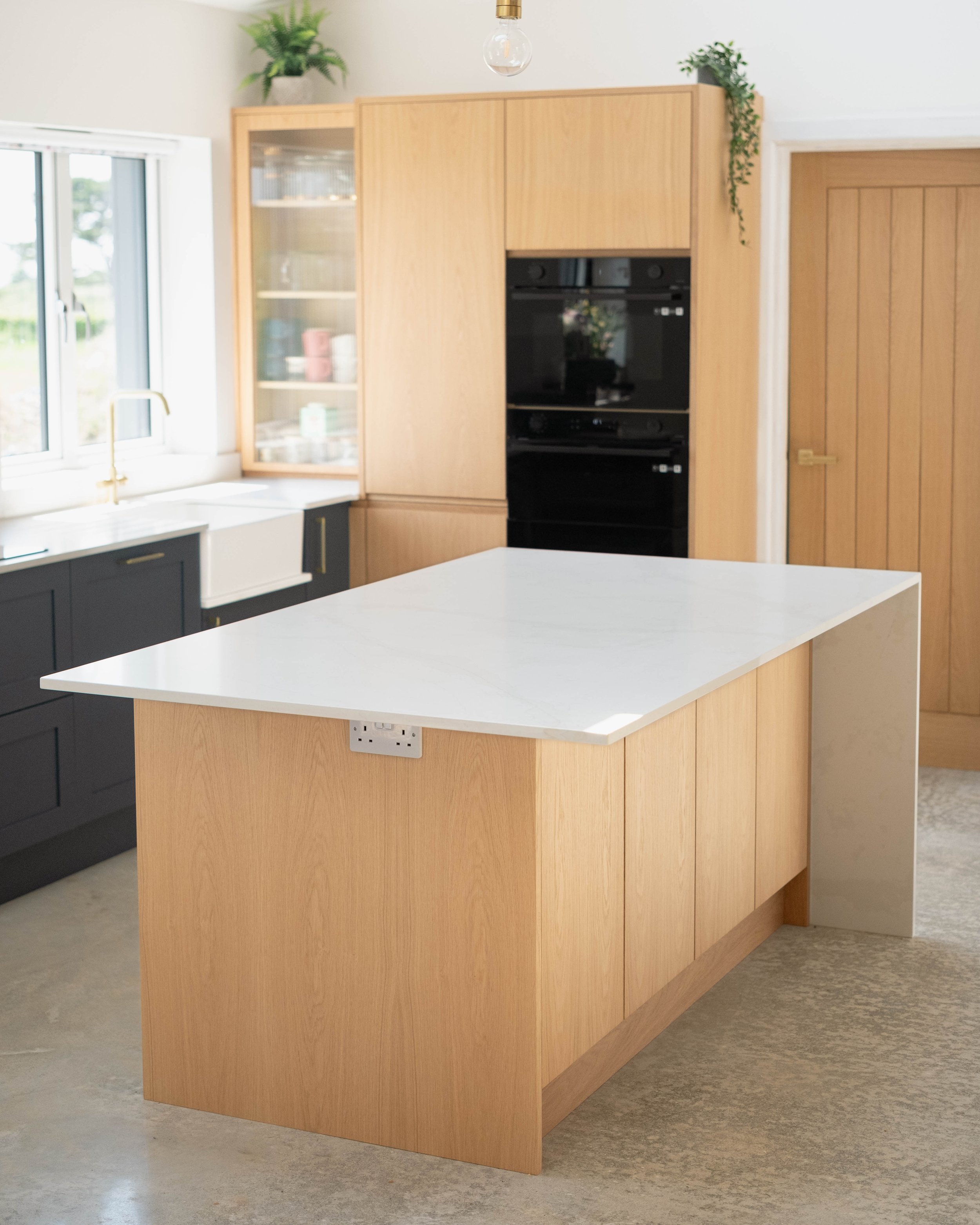 A modern kitchen with a white countertop island, dark cabinets below, large window with a view outside, a gold faucet, light wood cabinets, a black built-in oven, and green potted plants on top of the cabinets.
