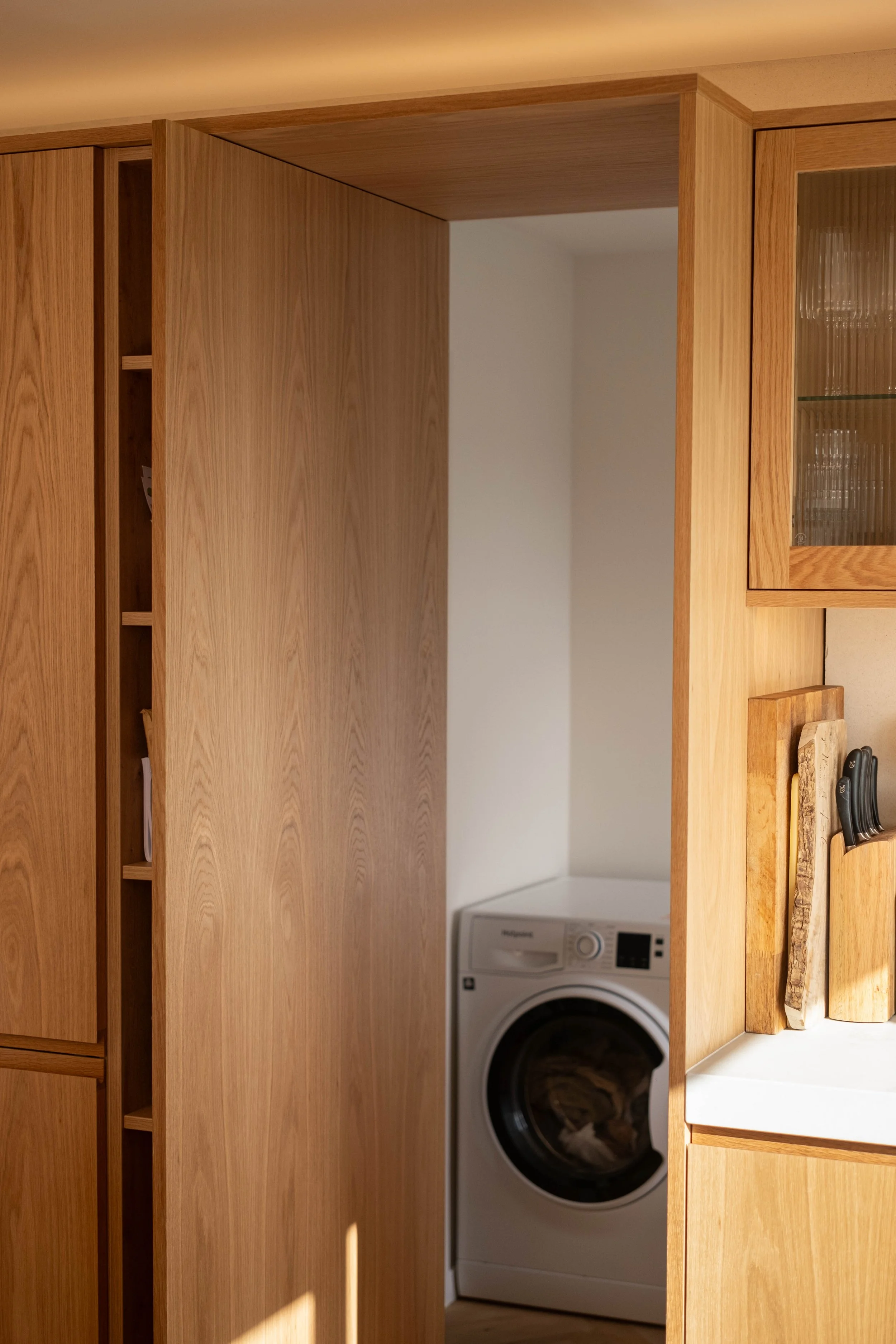 A laundry room with wooden cabinets and a washing machine. The cabinets have glass doors, and there is a wooden countertop with a loaf of bread and some utensils on it.
