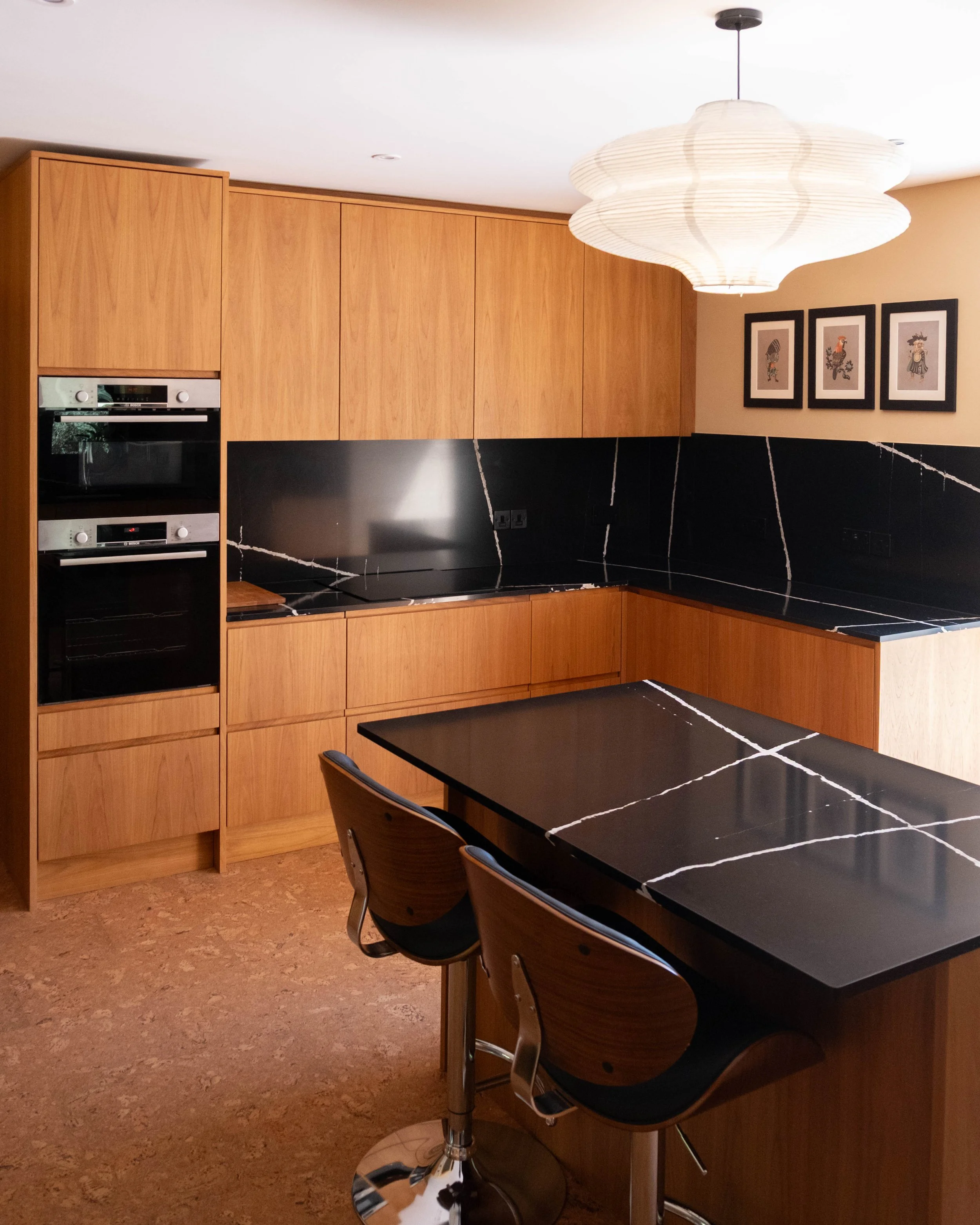 A modern kitchen with wooden cabinets, a black marble counter, and a black dining table with chairs in front. A large paper lantern hangs from the ceiling, and three framed art pieces are on the wall.