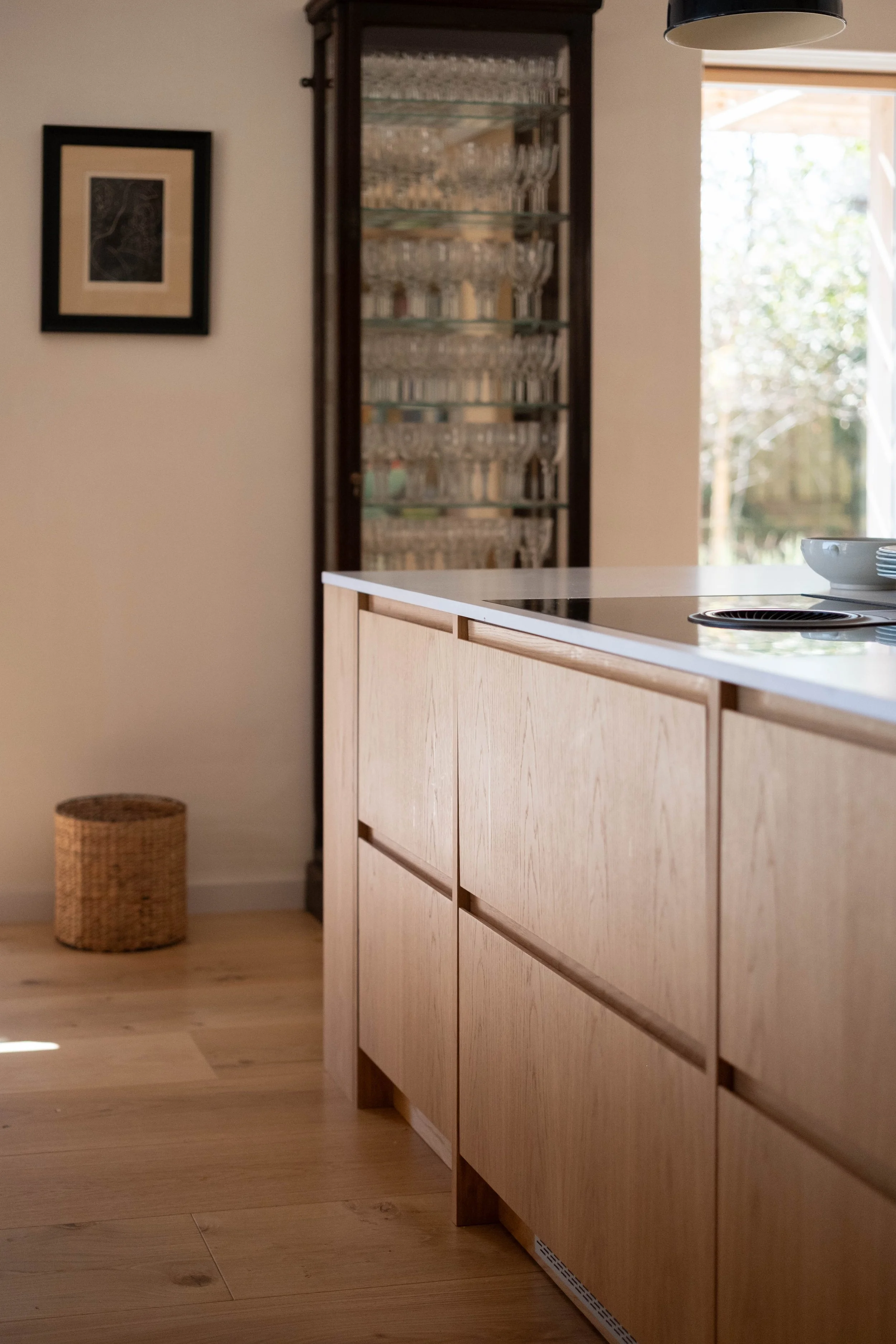 A modern kitchen with a wooden island, a glass-fronted cabinet filled with glassware, a framed artwork on the wall, a small wicker basket on the floor, and a large window allowing natural light to illuminate the space.