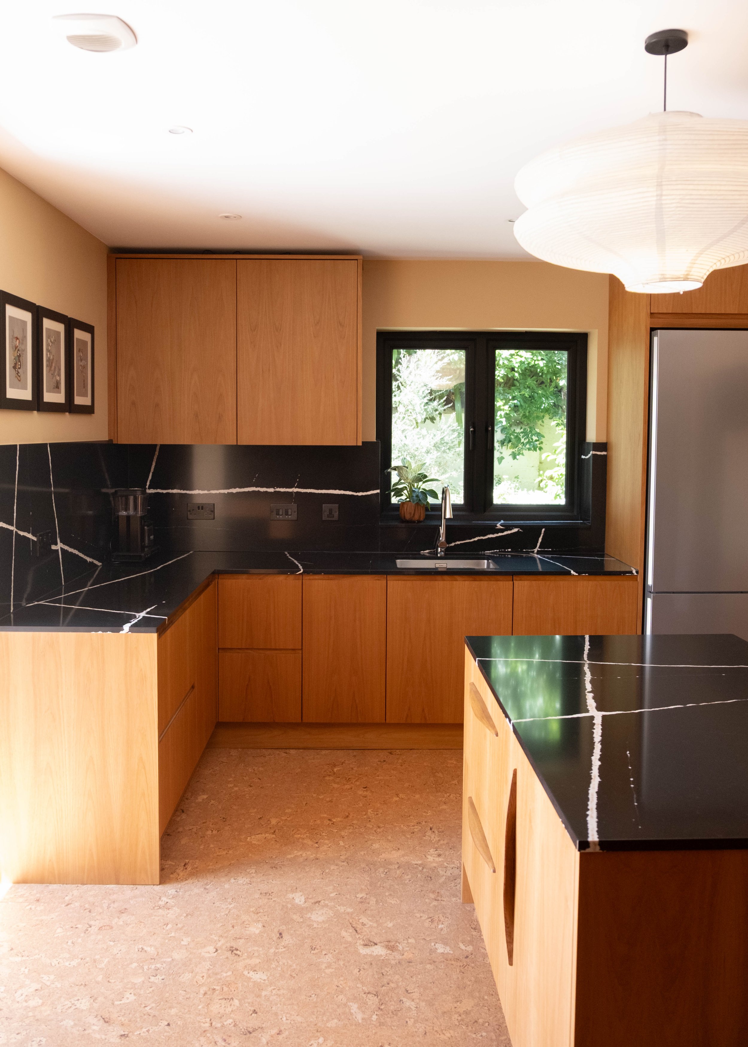 Modern kitchen with wooden cabinets, black marble countertops with white veining, a window with a view of greenery, and a ceiling pendant light.