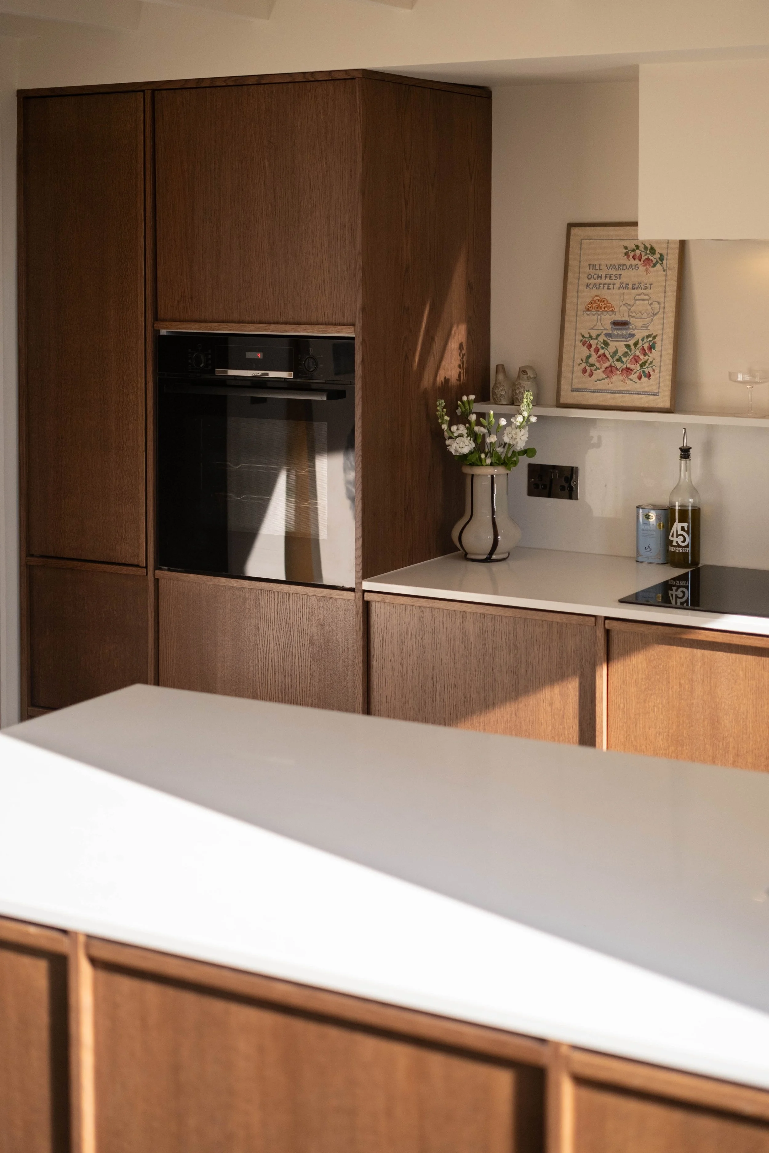 A kitchen with wooden cabinets, a white countertop, and a shelf with decorative items including a framed cross-stitch picture, vases, a bottle, and a can. There is a flower vase with white flowers on the counter.