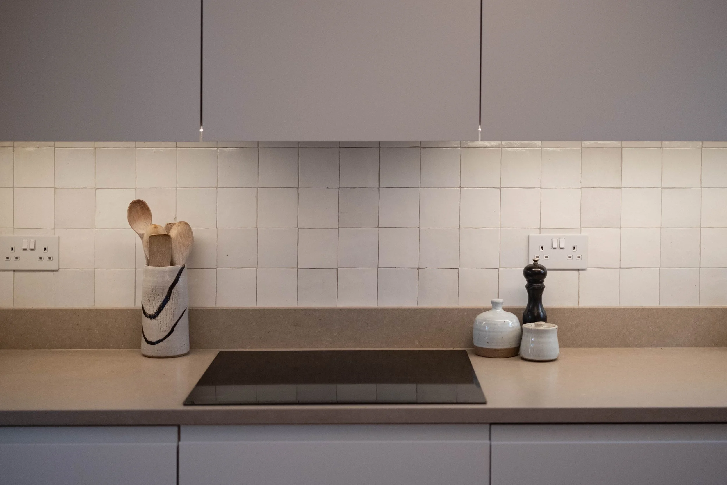 Kitchen countertop with a black induction stove, a container holding wooden utensils, and three ceramic vases in different sizes and colors, against a tiled wall with three electrical outlets.