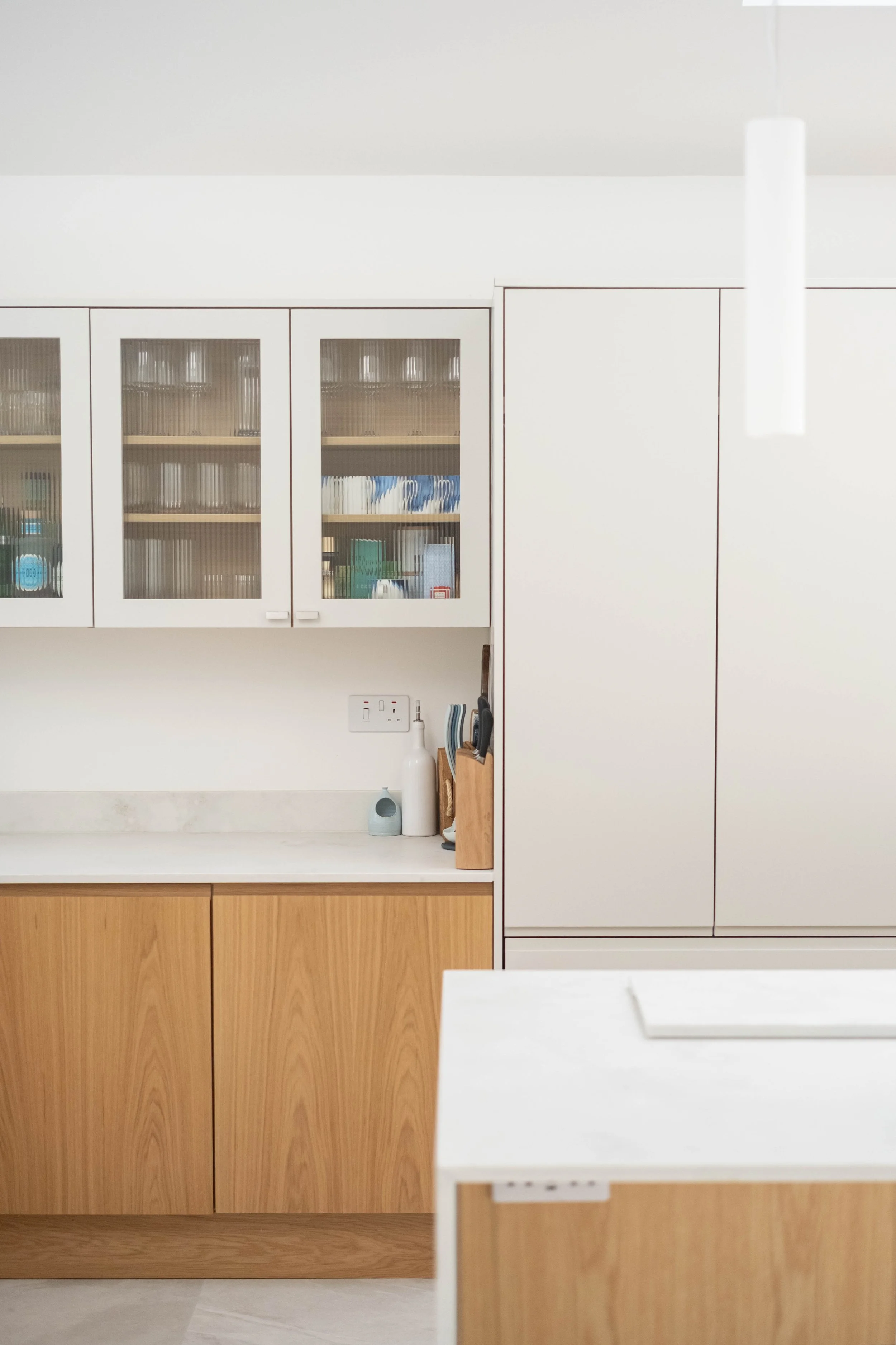 A modern kitchen with white upper cabinets with glass doors and a wooden lower cabinet. Items like books, a blue object, and a red box are visible inside the cabinet. On the countertop, there are kitchen utensils, a paper towel roll, and a white pitc