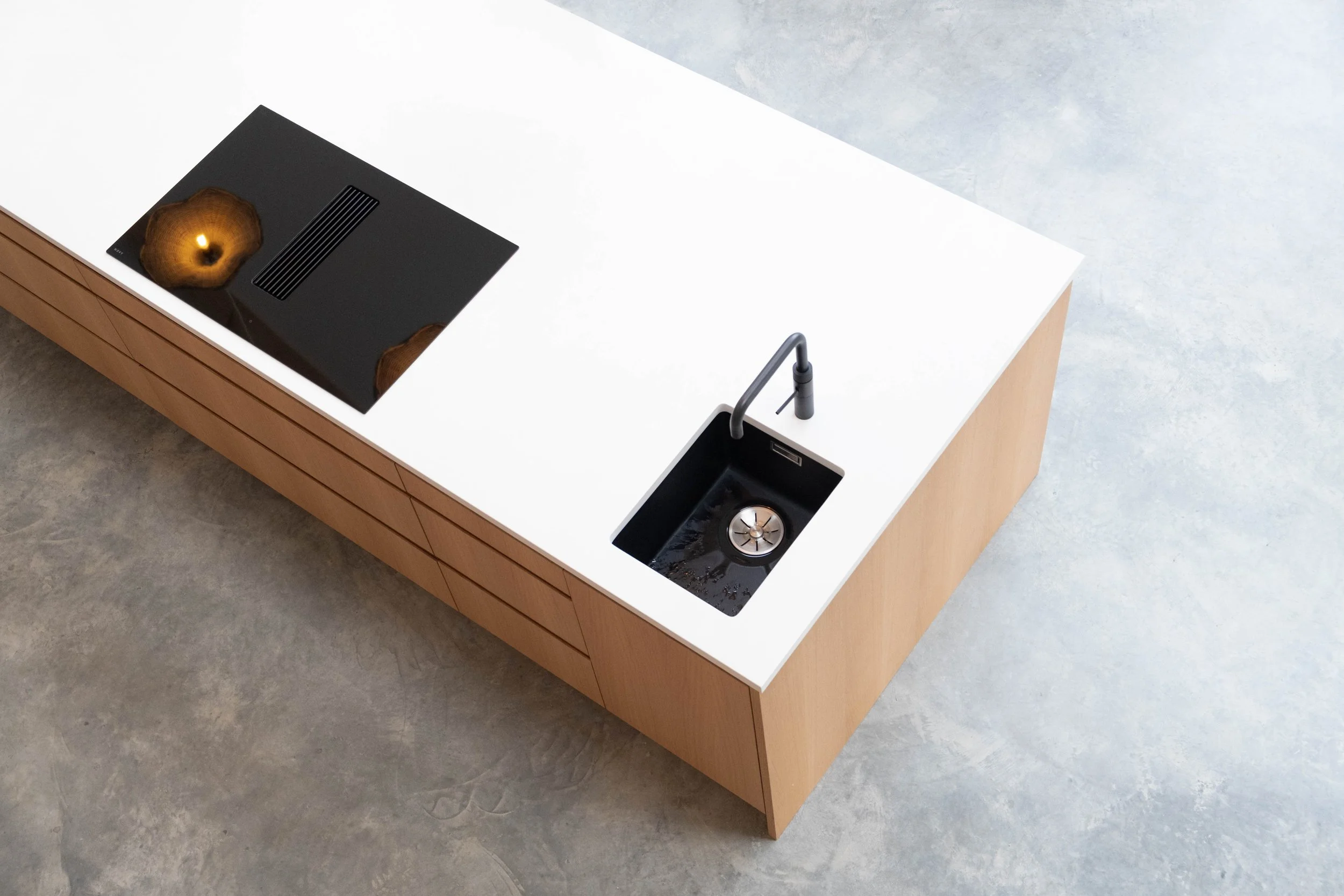 Modern kitchen with white countertop, black induction cooktop with a reflection, and a black sink with a silver faucet. Wooden cabinetry front panels and a concrete floor.