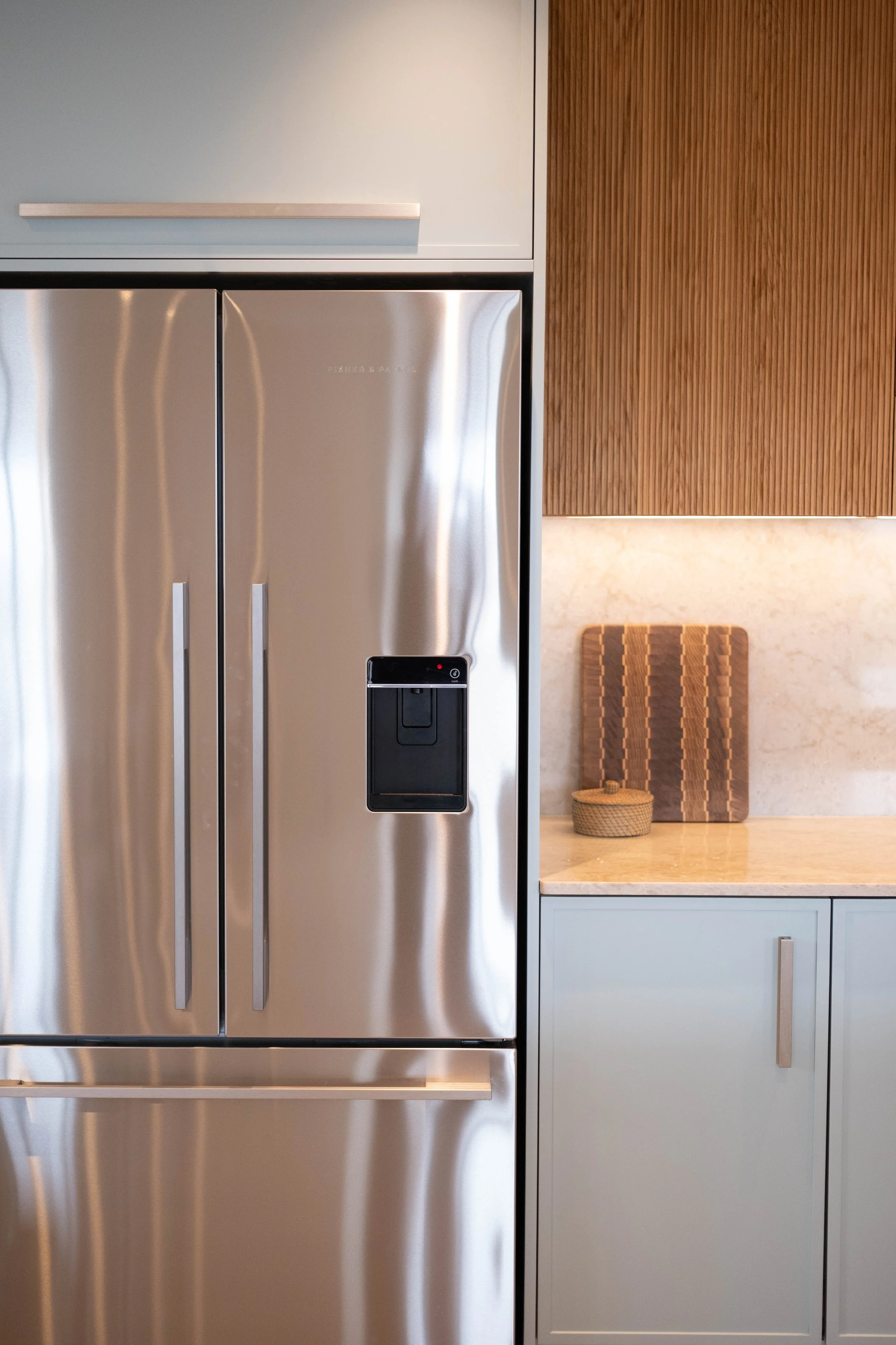 A modern kitchen with a stainless steel refrigerator, light blue cabinets with wooden handles, a beige marble countertop, and a wooden chair with a wicker basket on top.