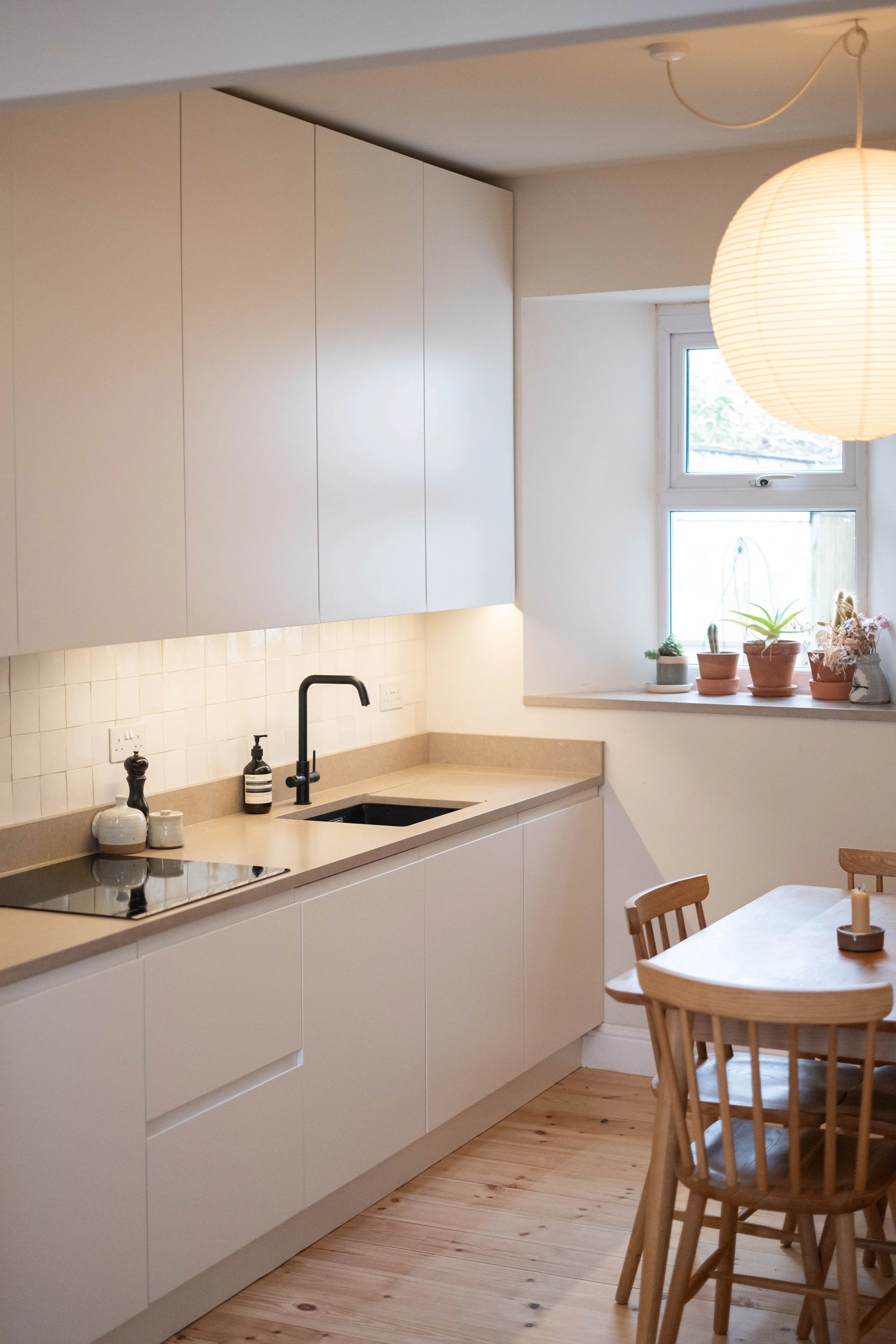 A minimalist kitchen with light-colored cabinets, a beige countertop, and a black sink. The window above the countertop has several potted plants on the sill, and a paper lantern hangs from the ceiling. A wooden dining table and chairs are in the for