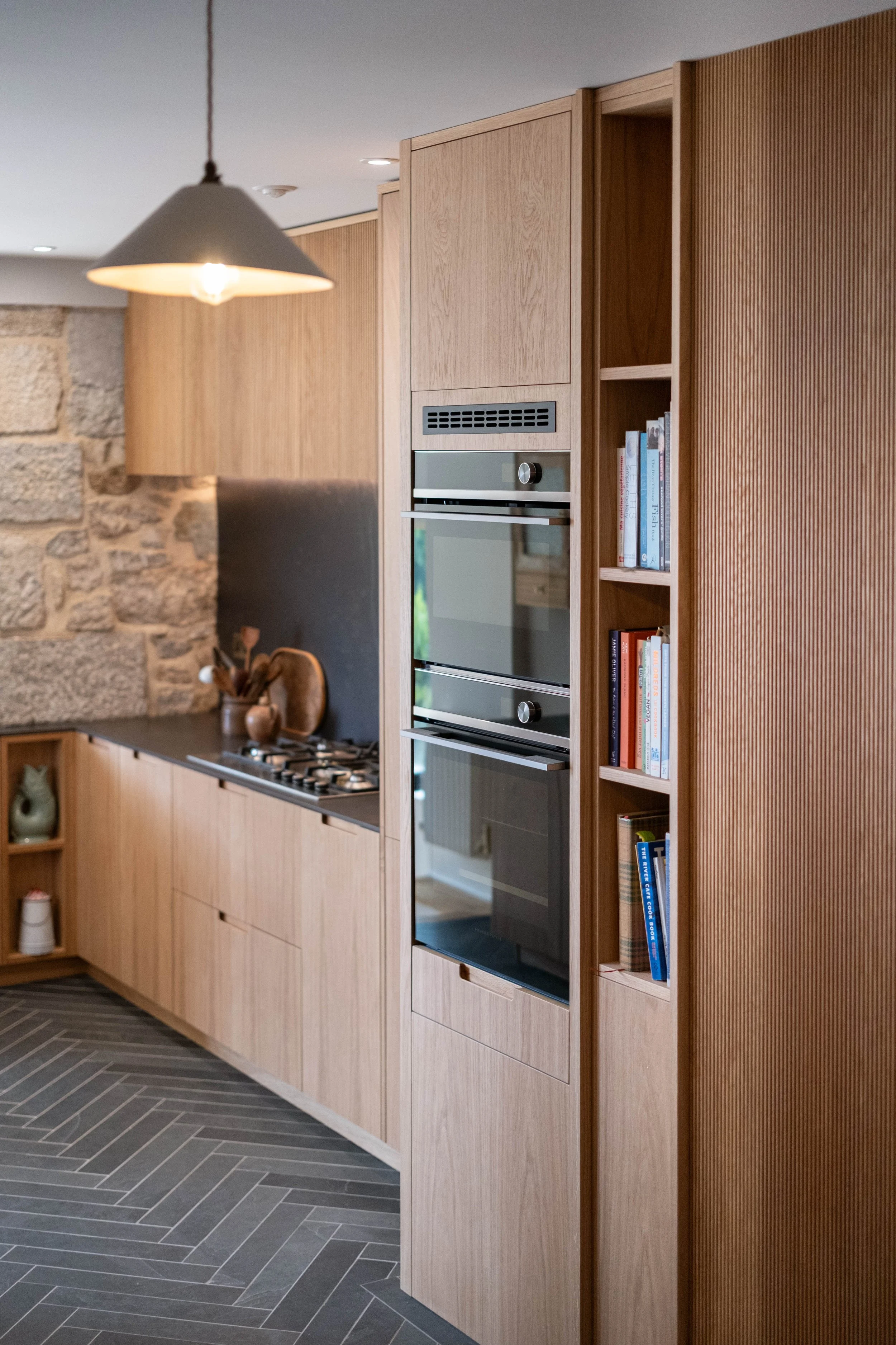 Modern kitchen with wood cabinets, built-in oven, bookshelf with cookbooks, stone wall, and gray herringbone tile floor, illuminated by a hanging pendant light.