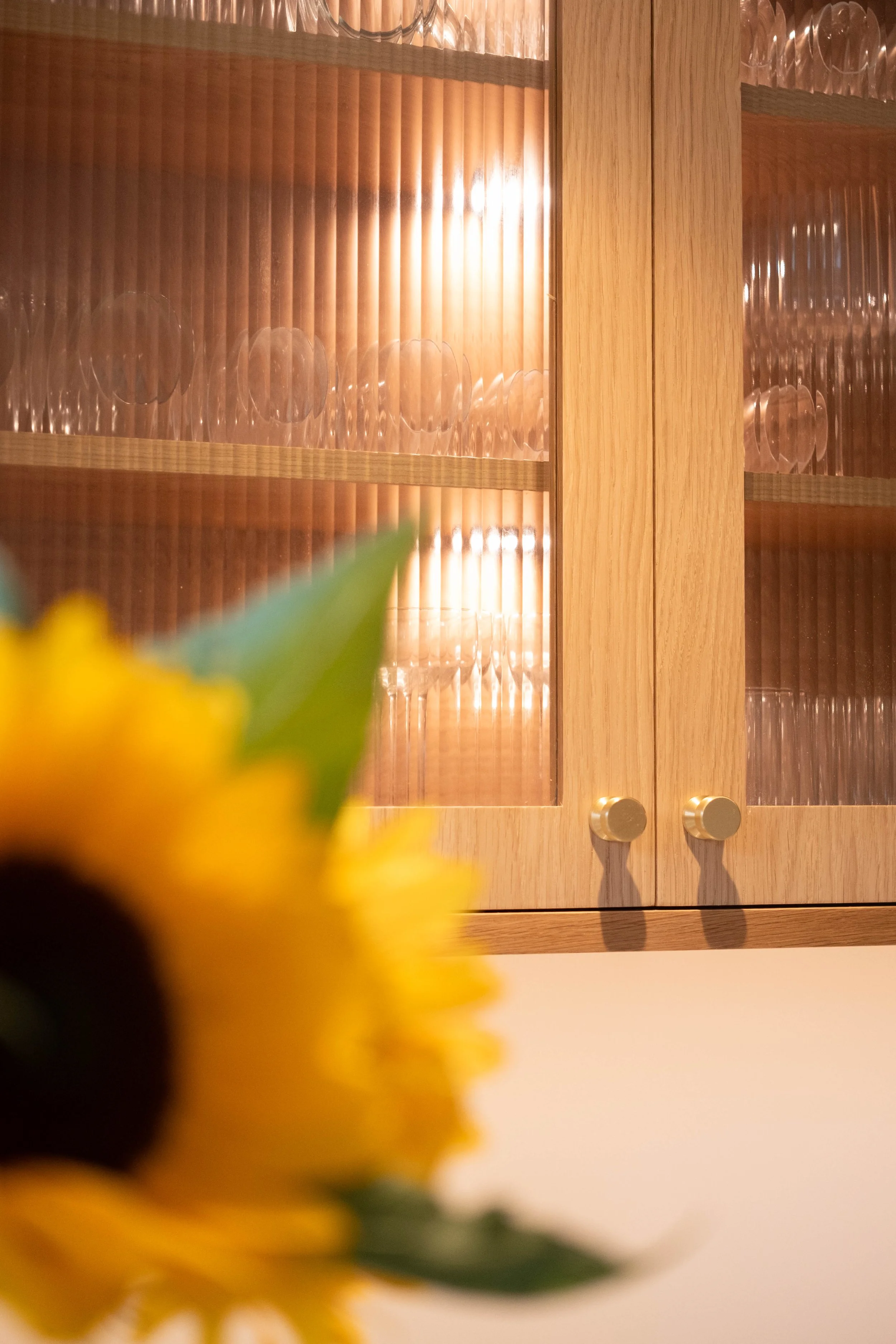 Blurred yellow flower in foreground with wooden cabinet with glass panels and round handles in the background.