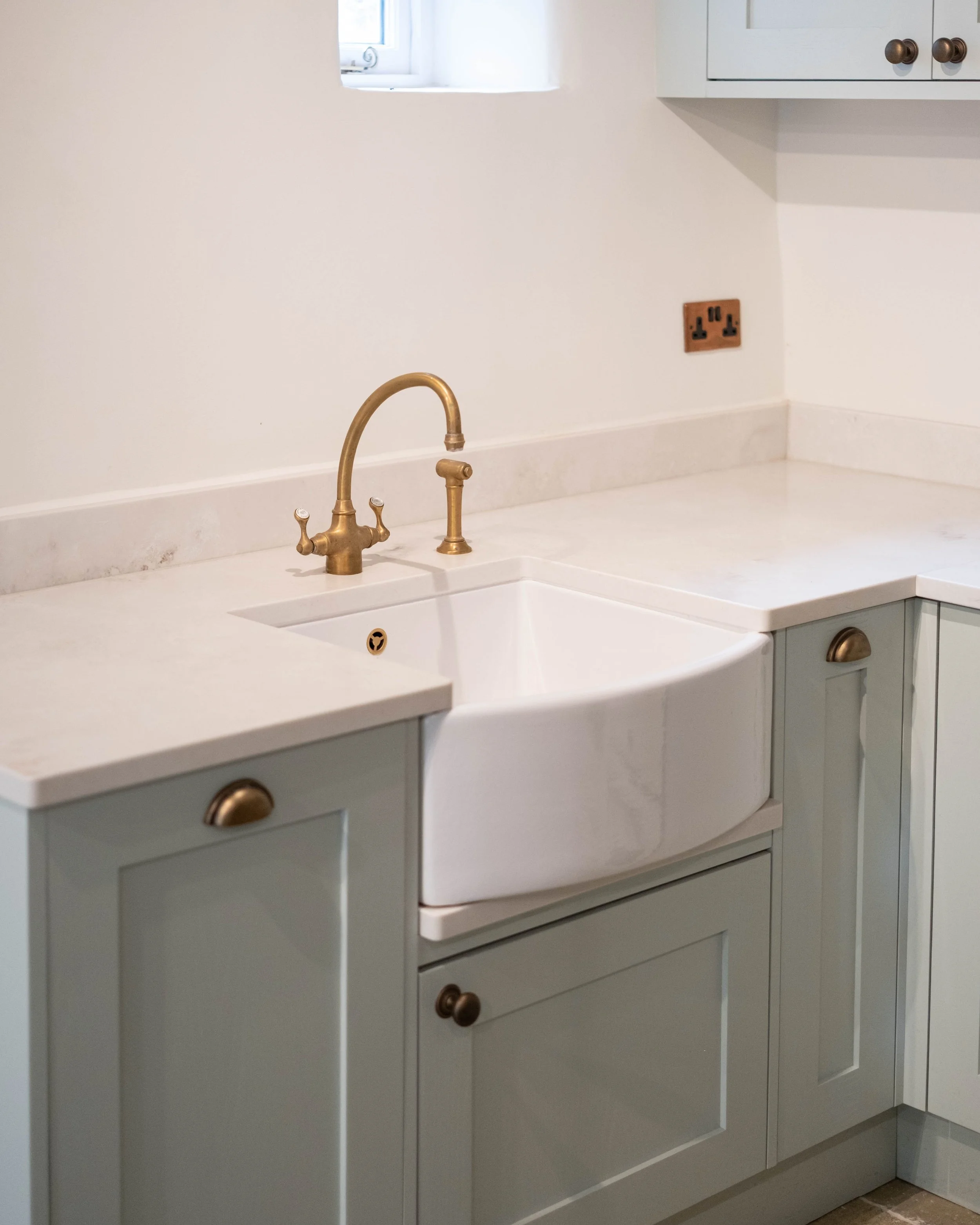 A kitchen countertop with a brass faucet and white sink, light green cabinets with round brass knobs, and a small window with a white frame above the sink.