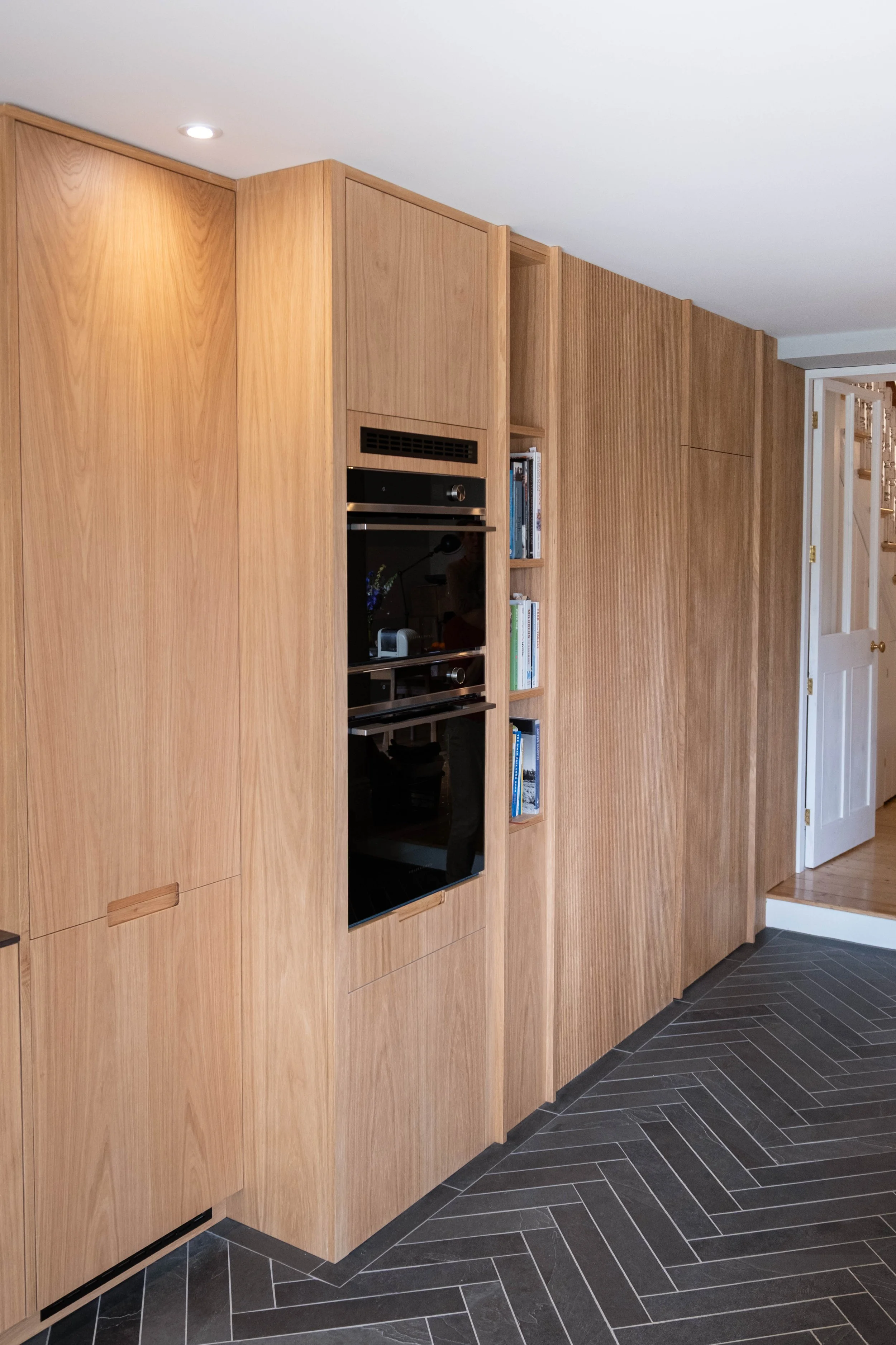 Wooden built-in kitchen cabinets with bookshelves and a black oven, situated on a black herringbone tile floor near a staircase.