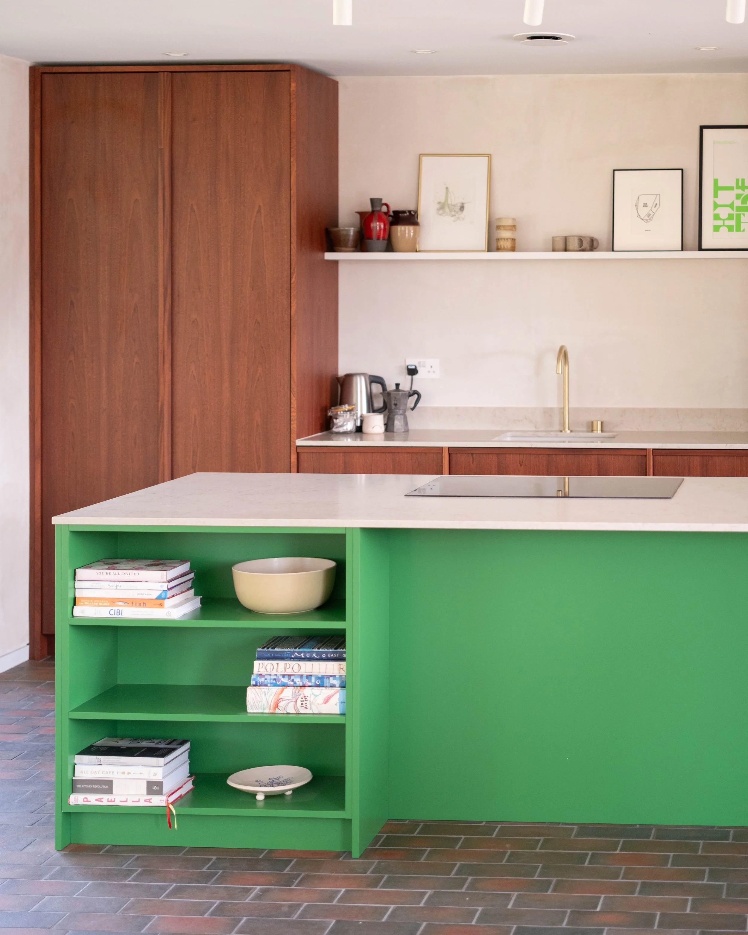 A kitchen with a green bookshelf, a marble countertop, a gold faucet, and a white wall with framed pictures and decorative items. There is a large wooden cabinet on the left and a tiled floor.
