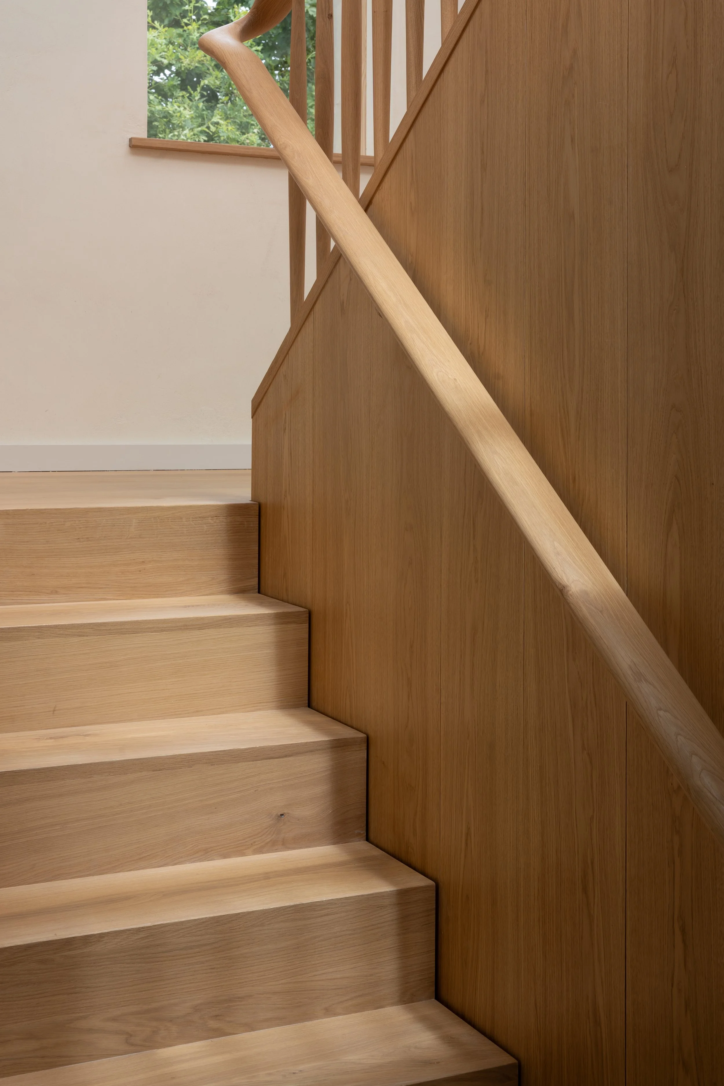 Wooden staircase with a handrail, next to a wall and a window showing green trees outside.