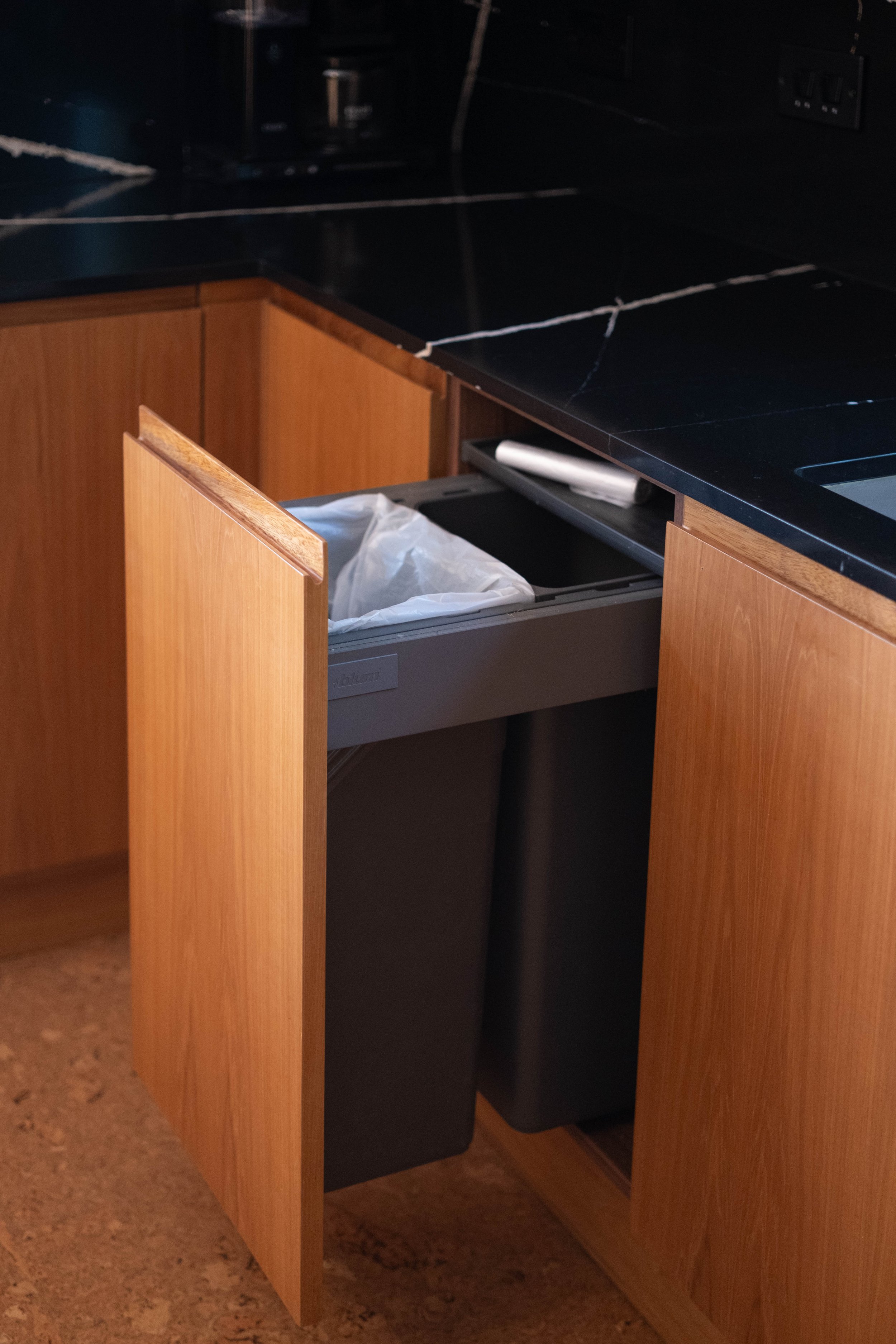 Open kitchen cabinet drawer with trash bag inside, against a black countertop and wooden cabinets.