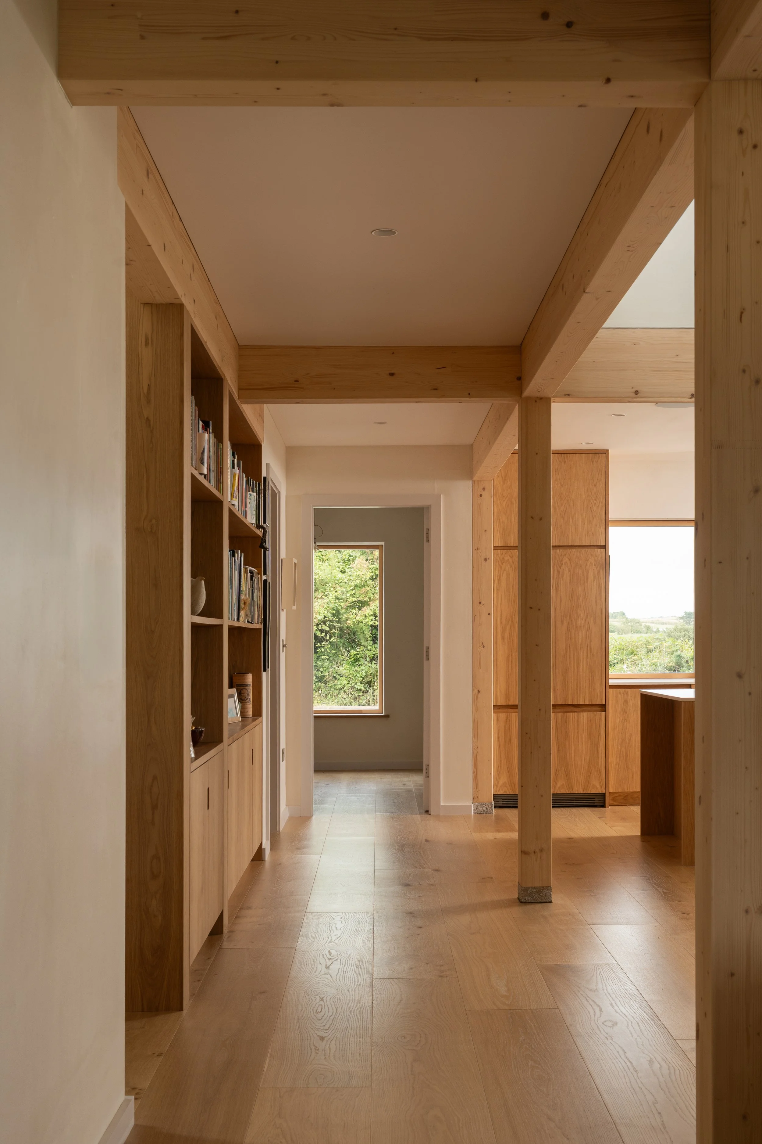 A hallway with wooden beams, a built-in bookshelf on the left, and windows letting in natural light in a modern home.