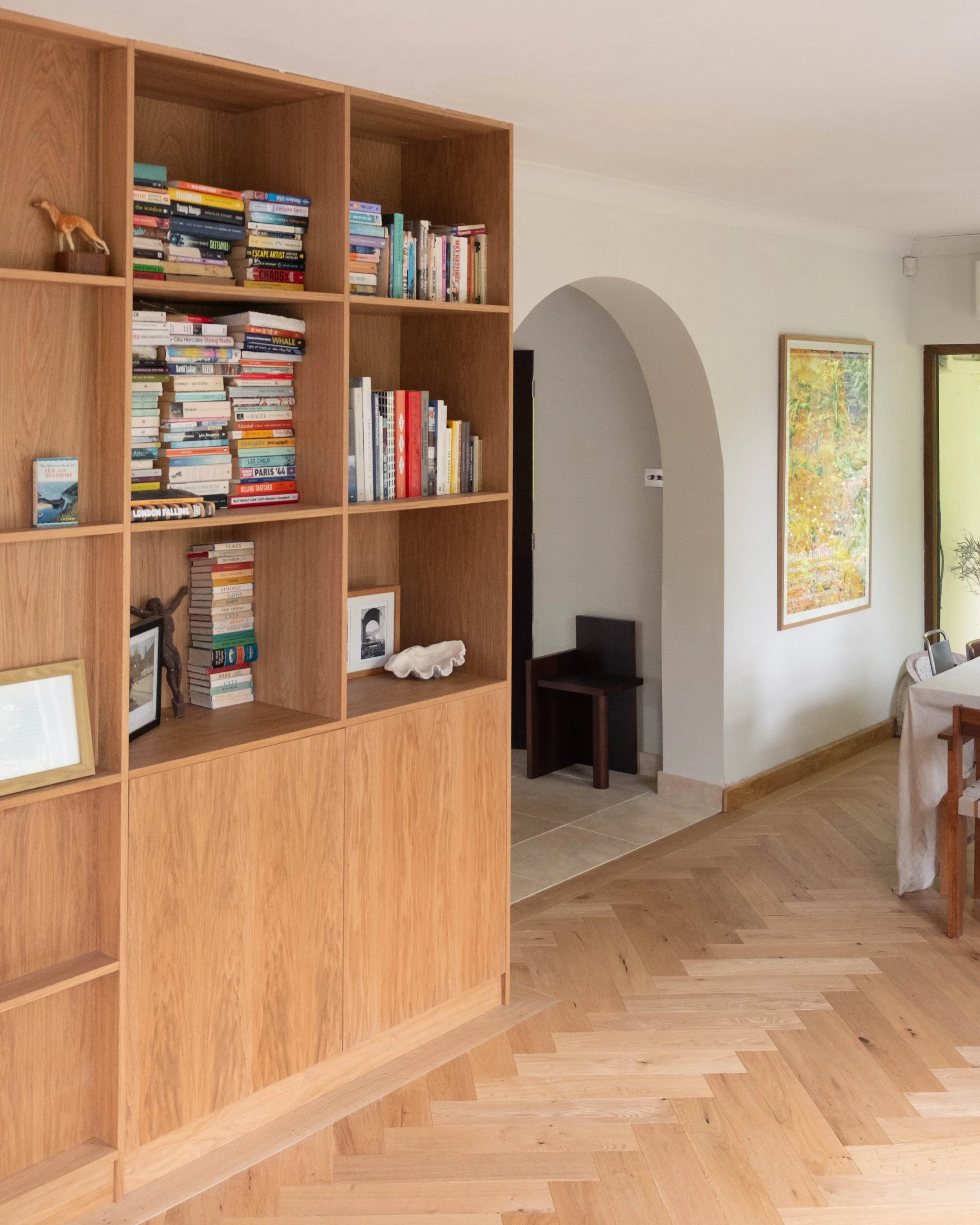 A wooden bookshelf filled with books and decorative items in a living room, with framed artwork on the wall and wooden flooring.