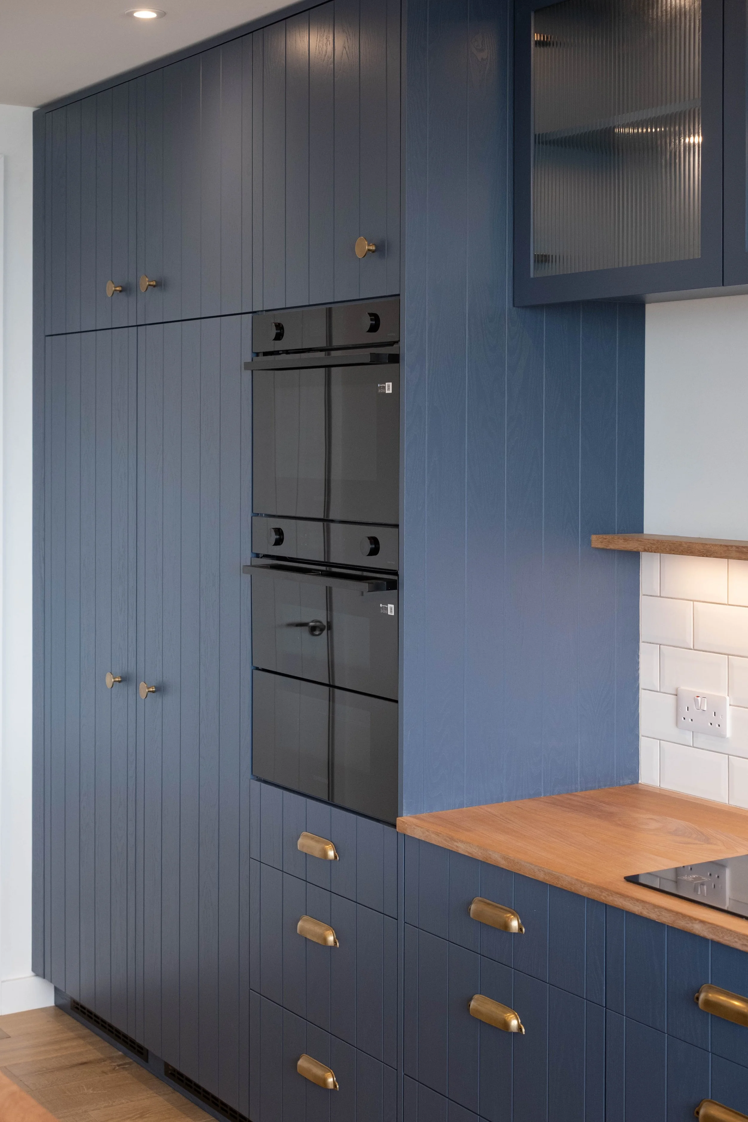 Blue kitchen cabinets with gold handles, a built-in black oven, white subway tile backsplash, a wooden countertop, and a white wall socket.
