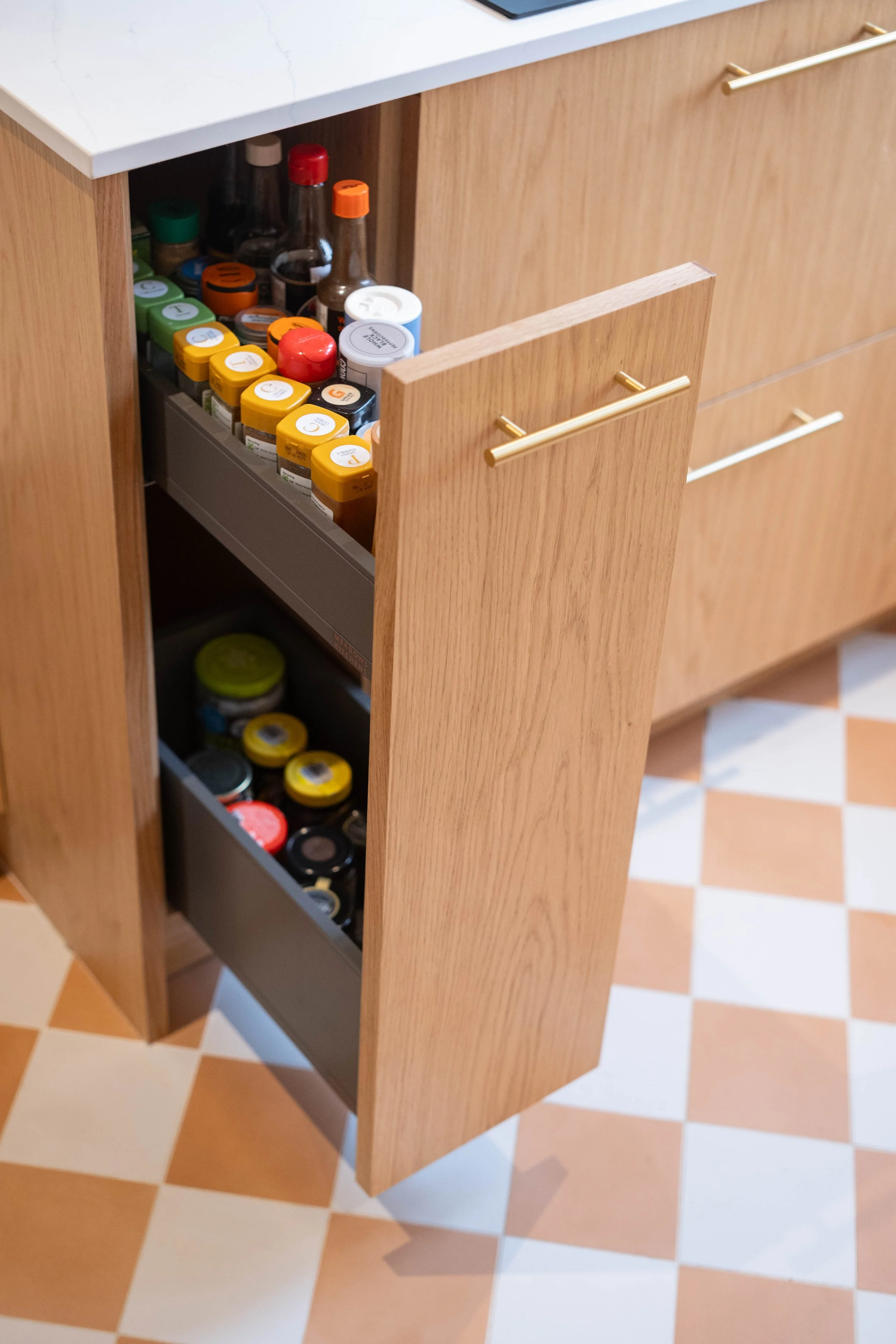 Open kitchen drawer with spice bottles and jars inside, against a wood cabinet and tiled floor.