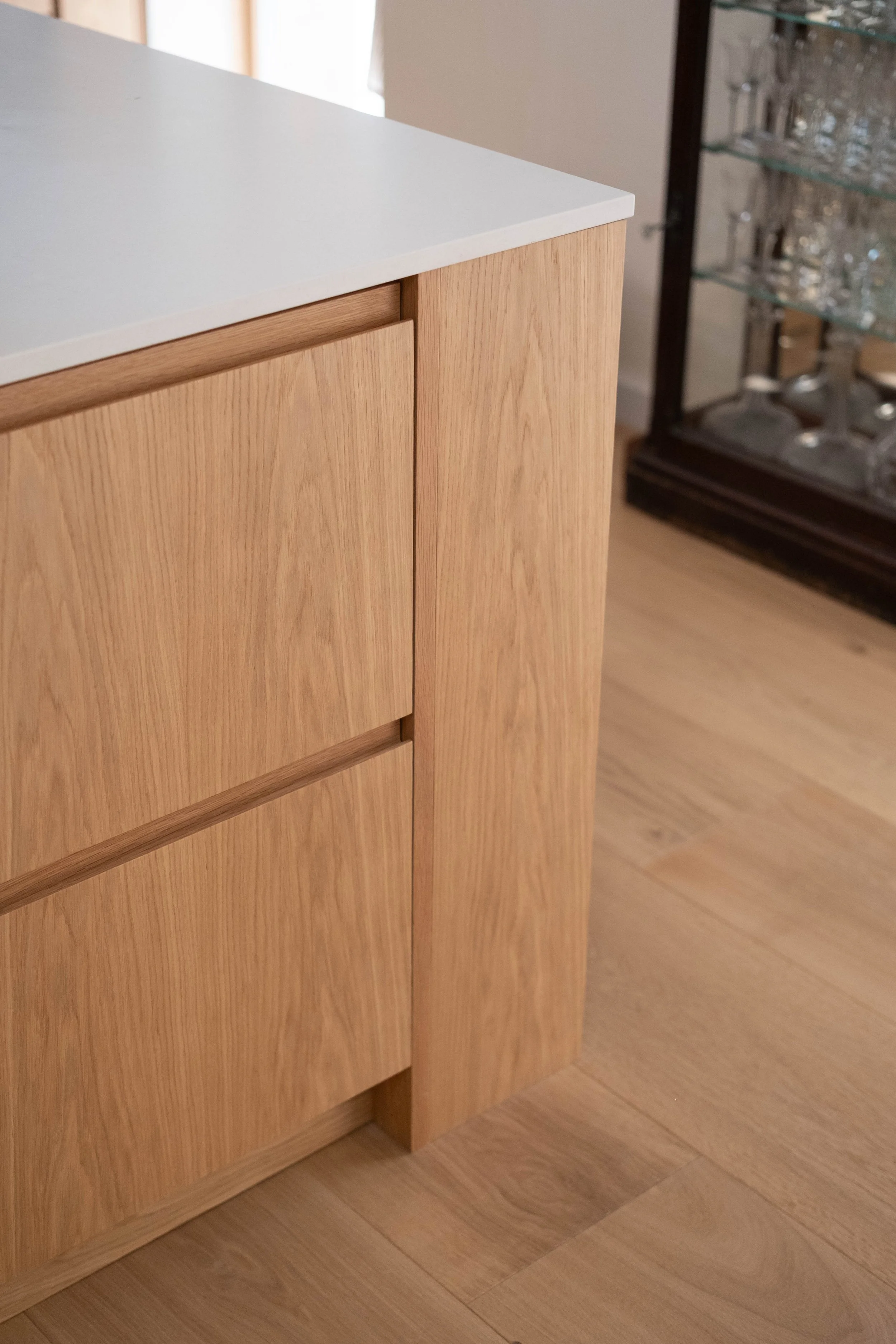 Close-up of a wooden cabinet with a light-colored countertop in a room with wooden flooring and a glass display cabinet with glassware in the background.