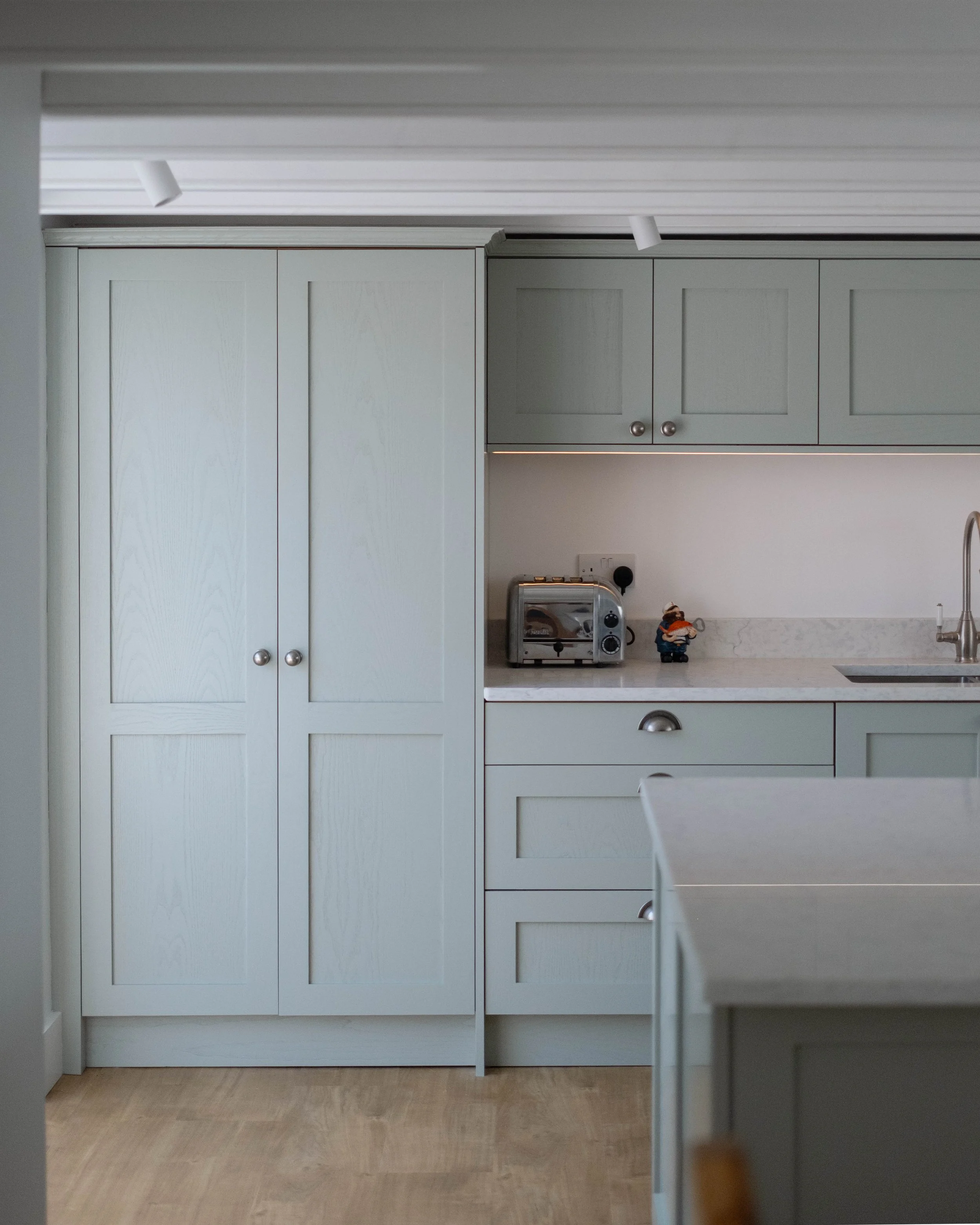 A kitchen with white cabinets, a white countertop, and a small kitchen appliance, possibly a toaster. There is a figure of a chef or chef doll on the counter. The image is taken from an angle that shows part of a dining table and flooring.