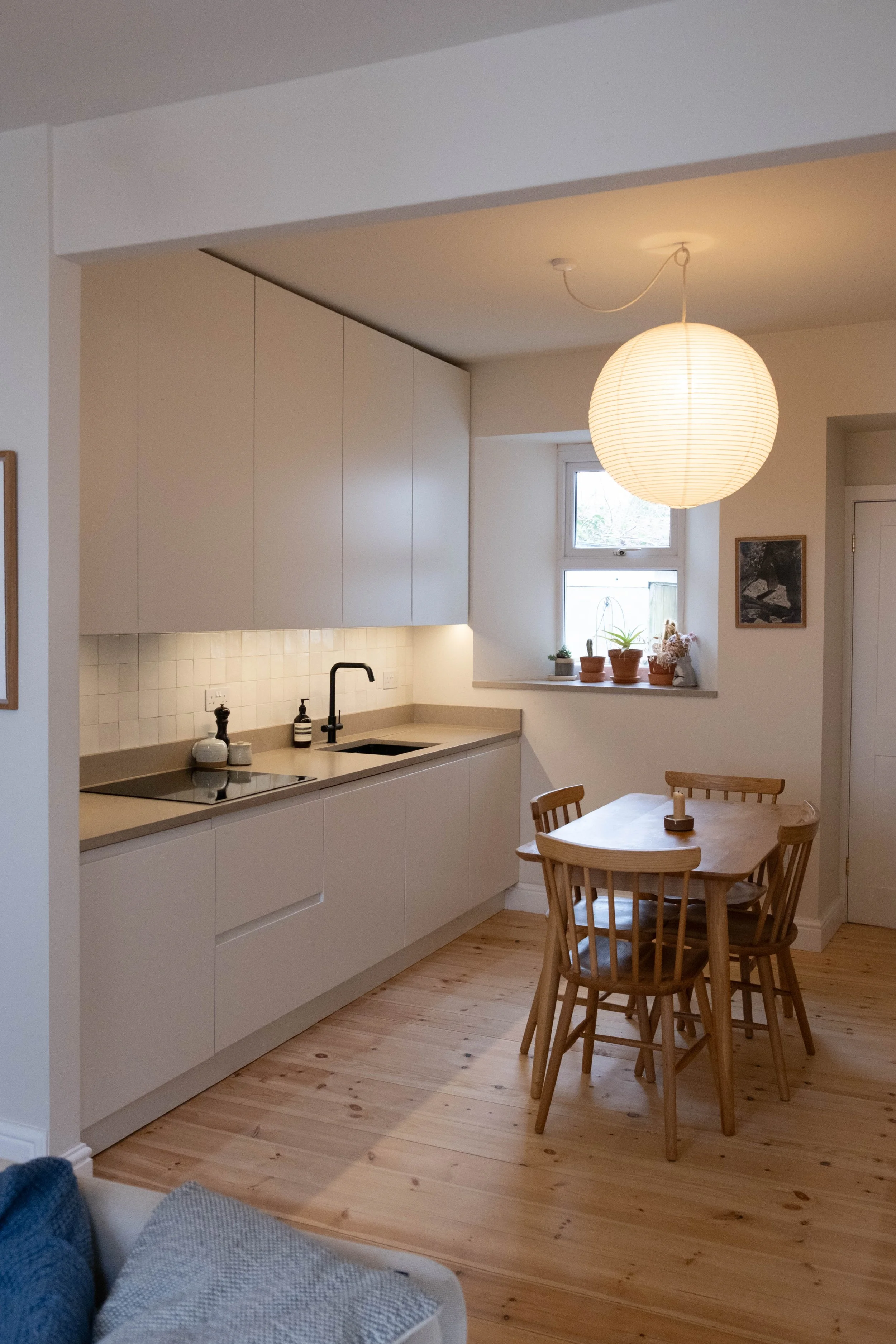 A modern kitchen with white cabinetry, a beige countertop, black faucet, and small potted plants on the windowsill. to the right, a wooden dining table with four chairs. There is a round paper lantern hanging from the ceiling, and a small window with