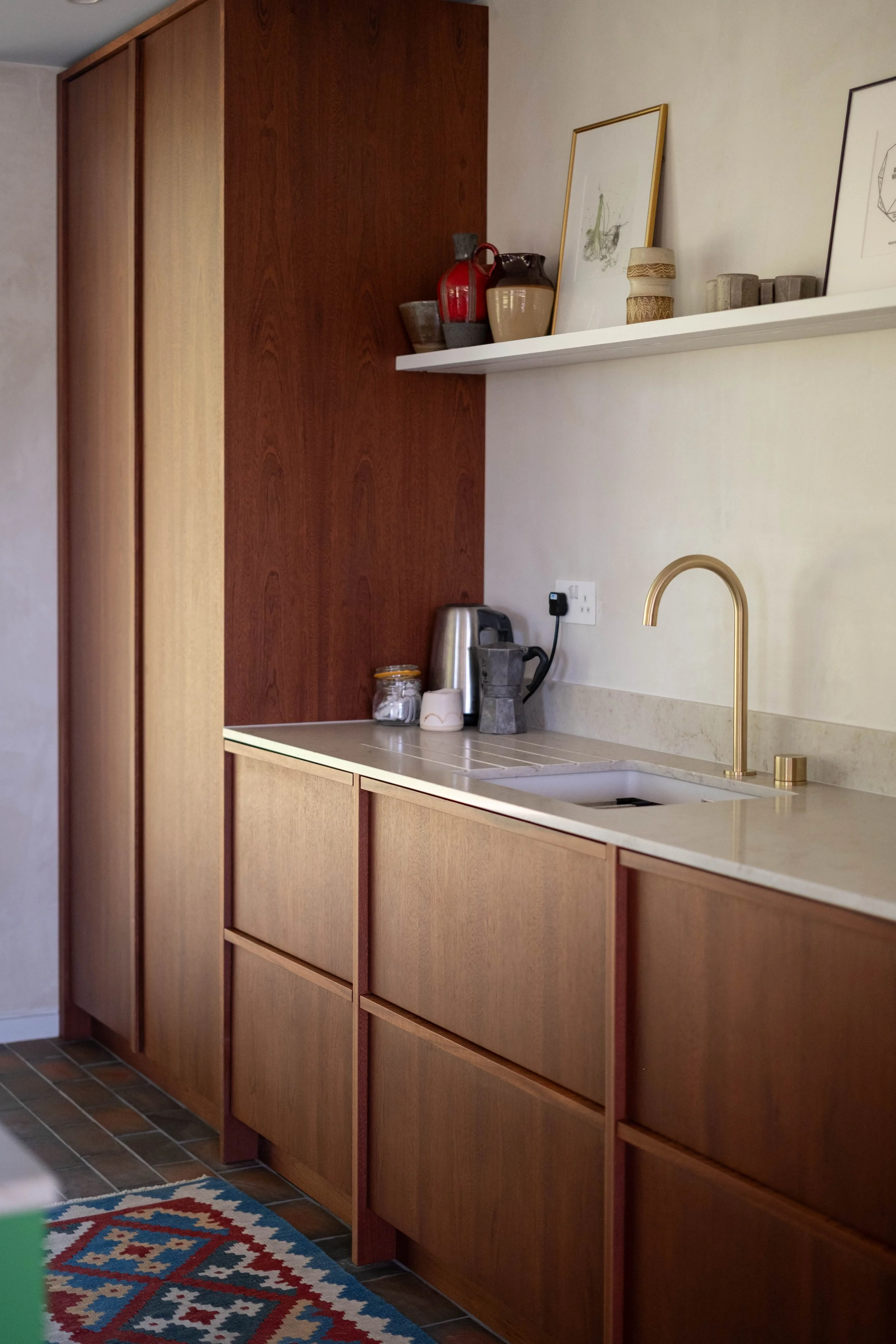 A kitchen with wooden cabinets, a beige countertop, and a white shelf with decorative items. There is a blue and red rug on the floor in front of the cabinets.