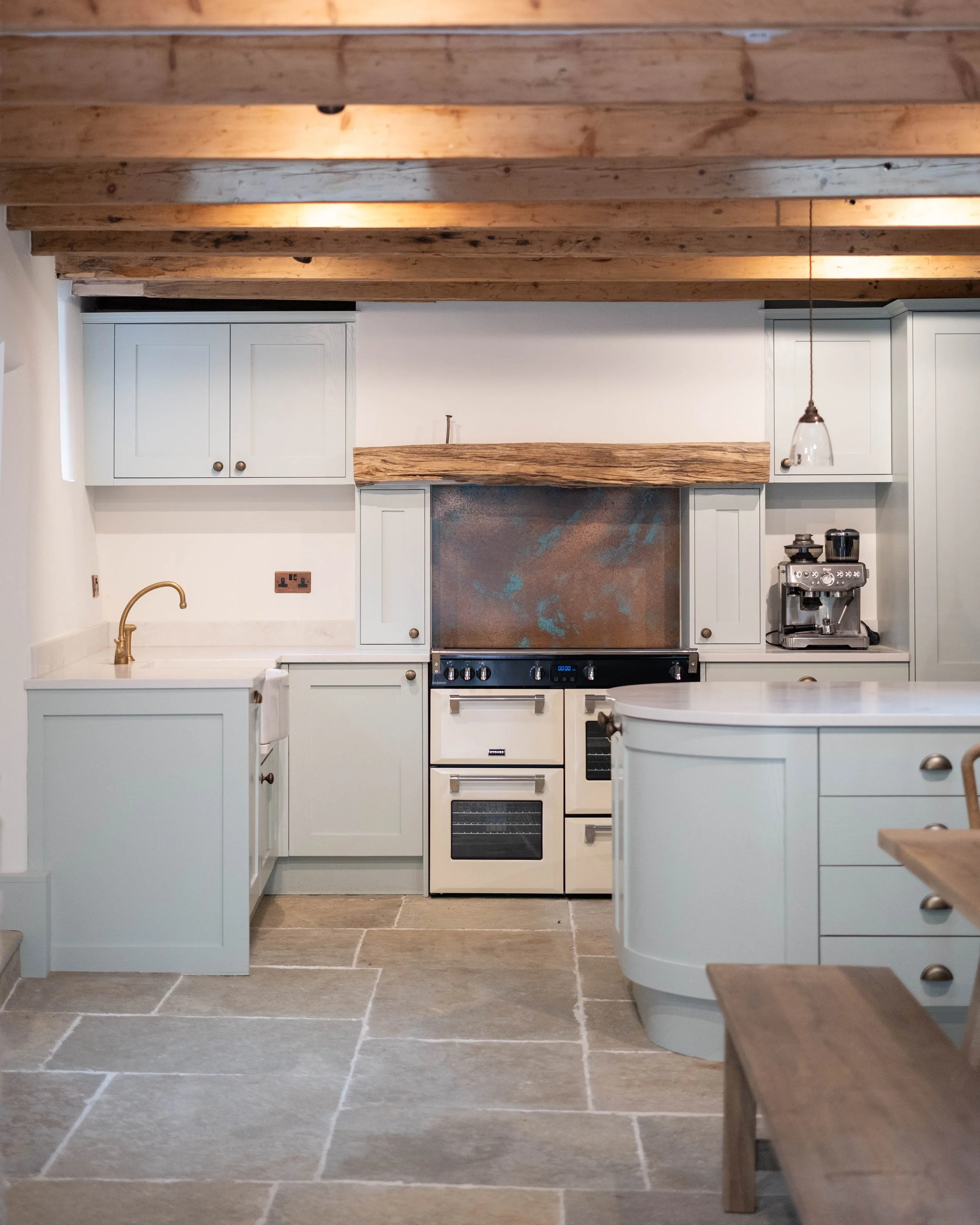 A cozy kitchen with white cabinets, a wooden beam above the stove, a coffee machine, and a gold faucet, with beige tiled flooring.