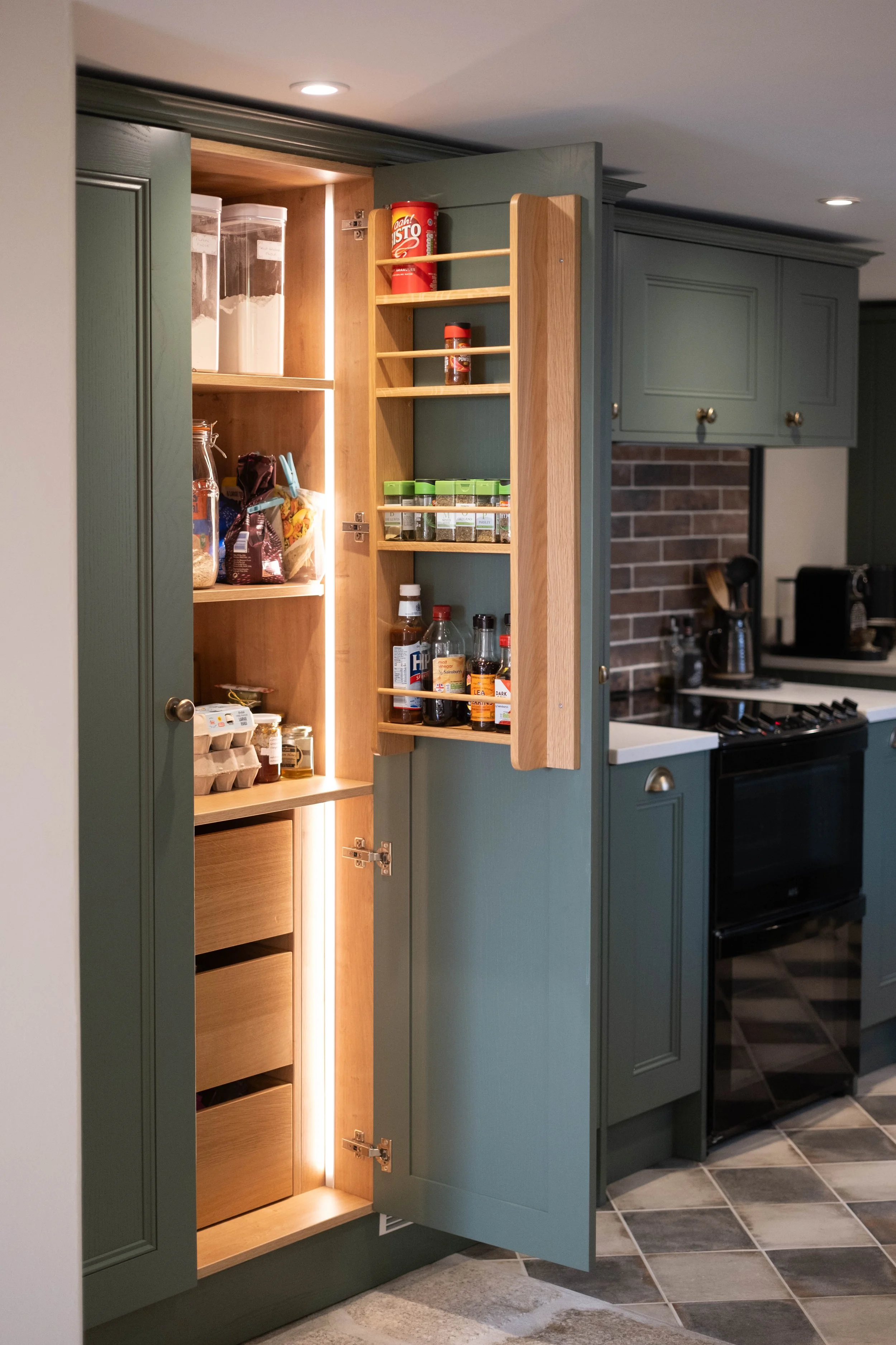 A kitchen cabinet with green exterior doors, one door open revealing a wooden interior with shelves holding various jars, bottles, and pantry items. The cabinet is located next to a kitchen area with tiled floor, a stove, and brick backsplash.