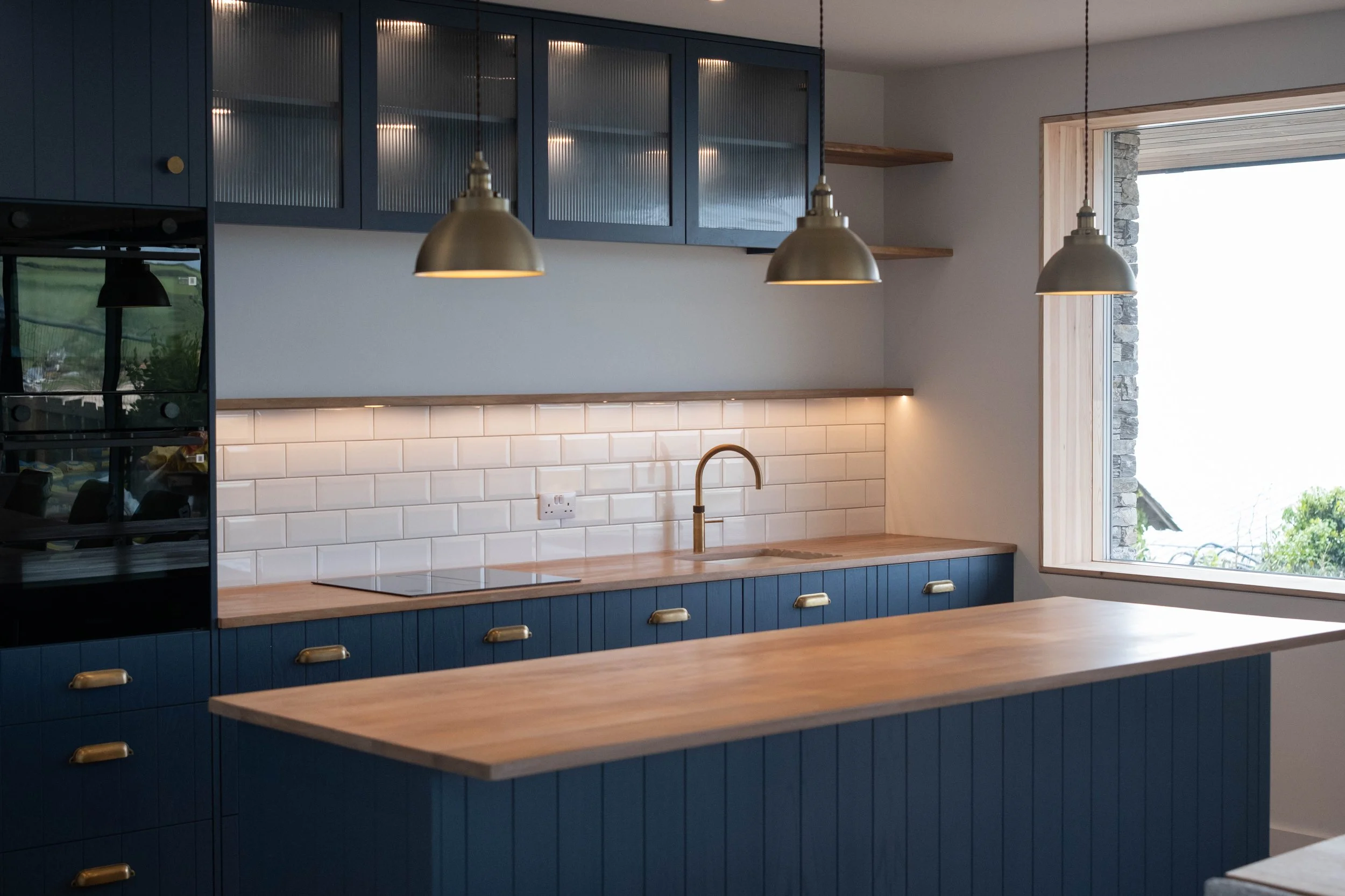 Modern kitchen with blue cabinetry, white subway tile backsplash, wooden countertops, and three hanging pendant lights. There is a large window showing an outdoor stone wall and greenery.