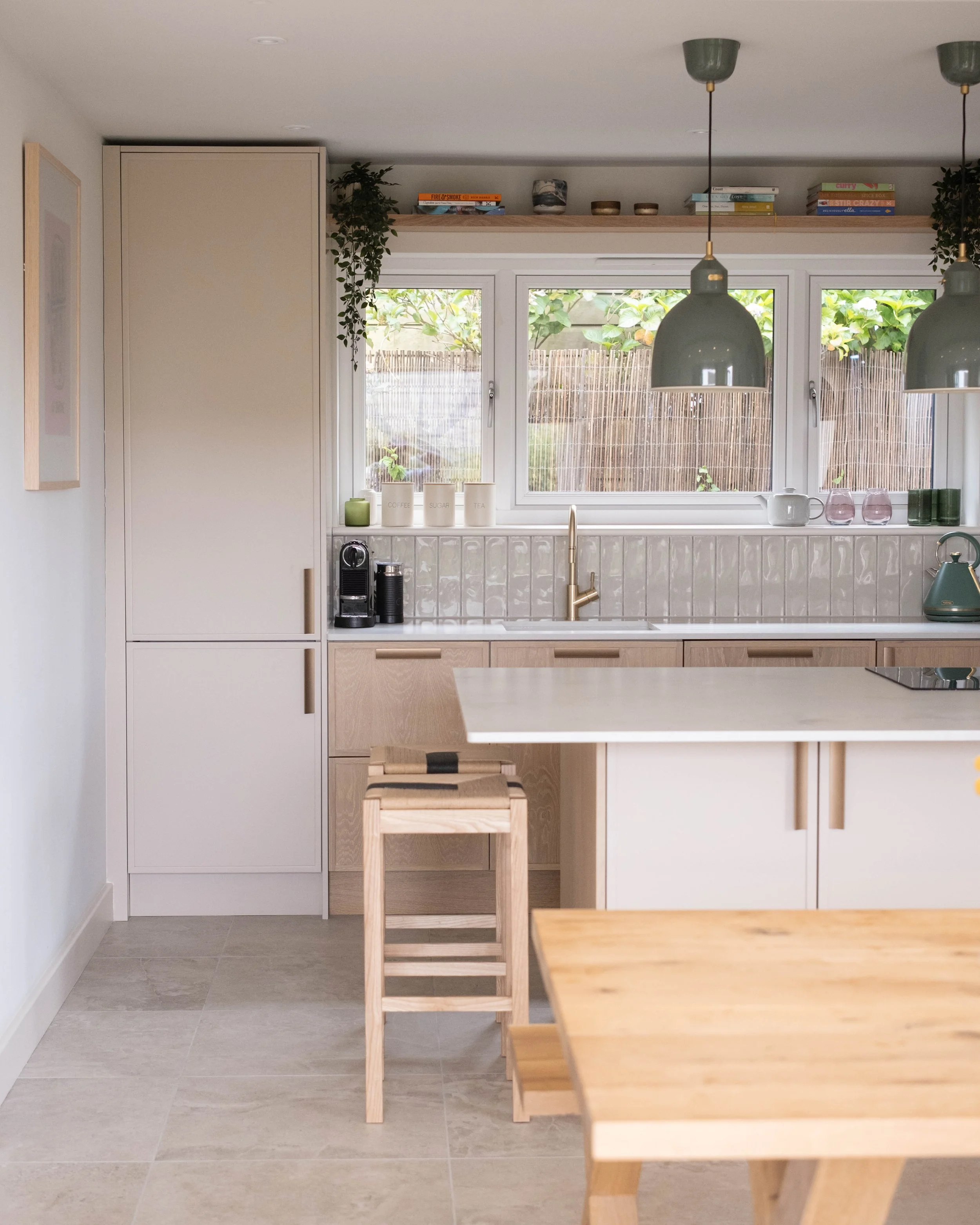 A minimalist kitchen with natural wood accents and white cabinetry, featuring a window overlooking a garden, pendant lighting, and a kitchen island with a light wood surface and stools.