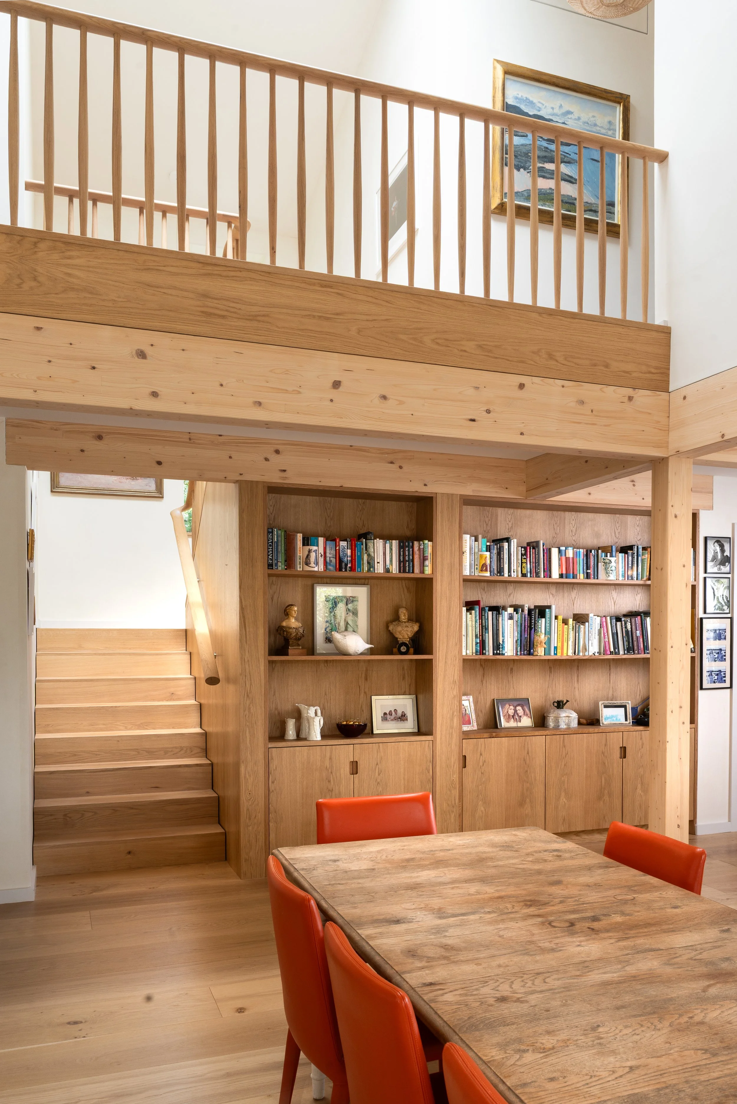 A cozy living room with a wooden built-in bookshelf filled with books and decorative items. There are stairs on the left side leading to a second level with a railing. A framed mountain landscape painting hangs on the wall. In the foreground, a woode
