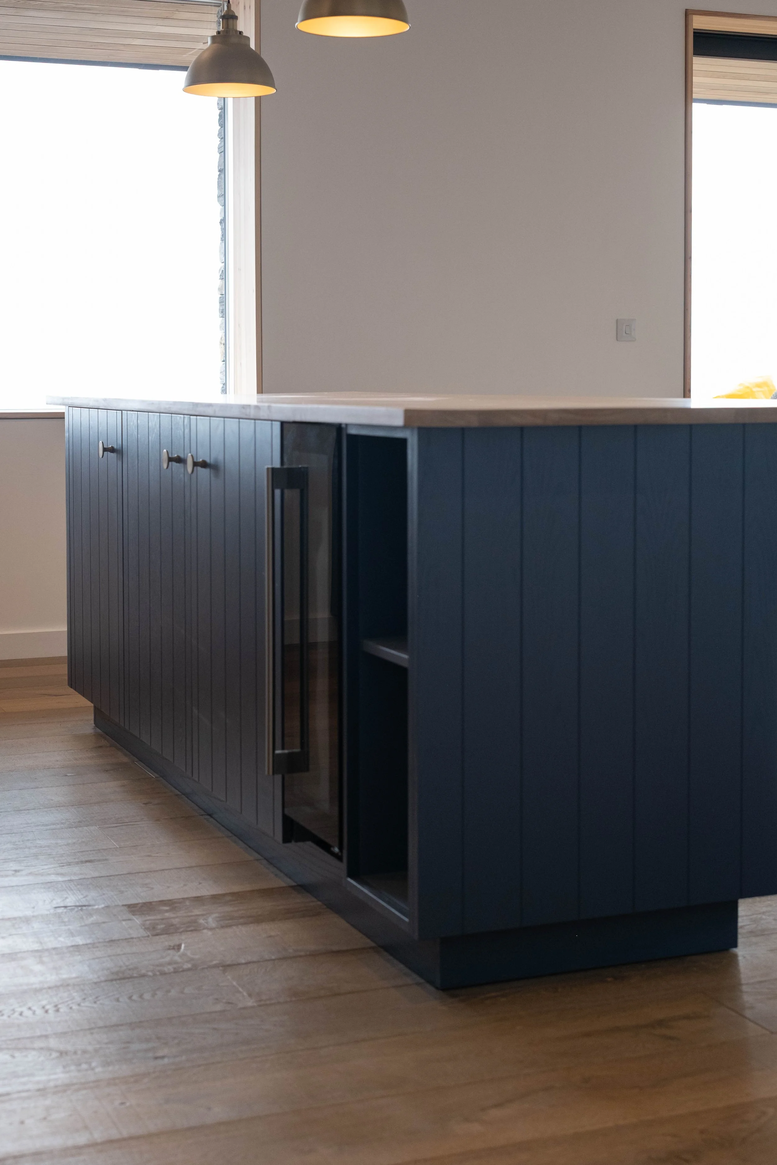 Modern kitchen island with blue vertical paneling, silver handles, a light wood countertop, and black base, situated on a wooden floor with large windows in background.