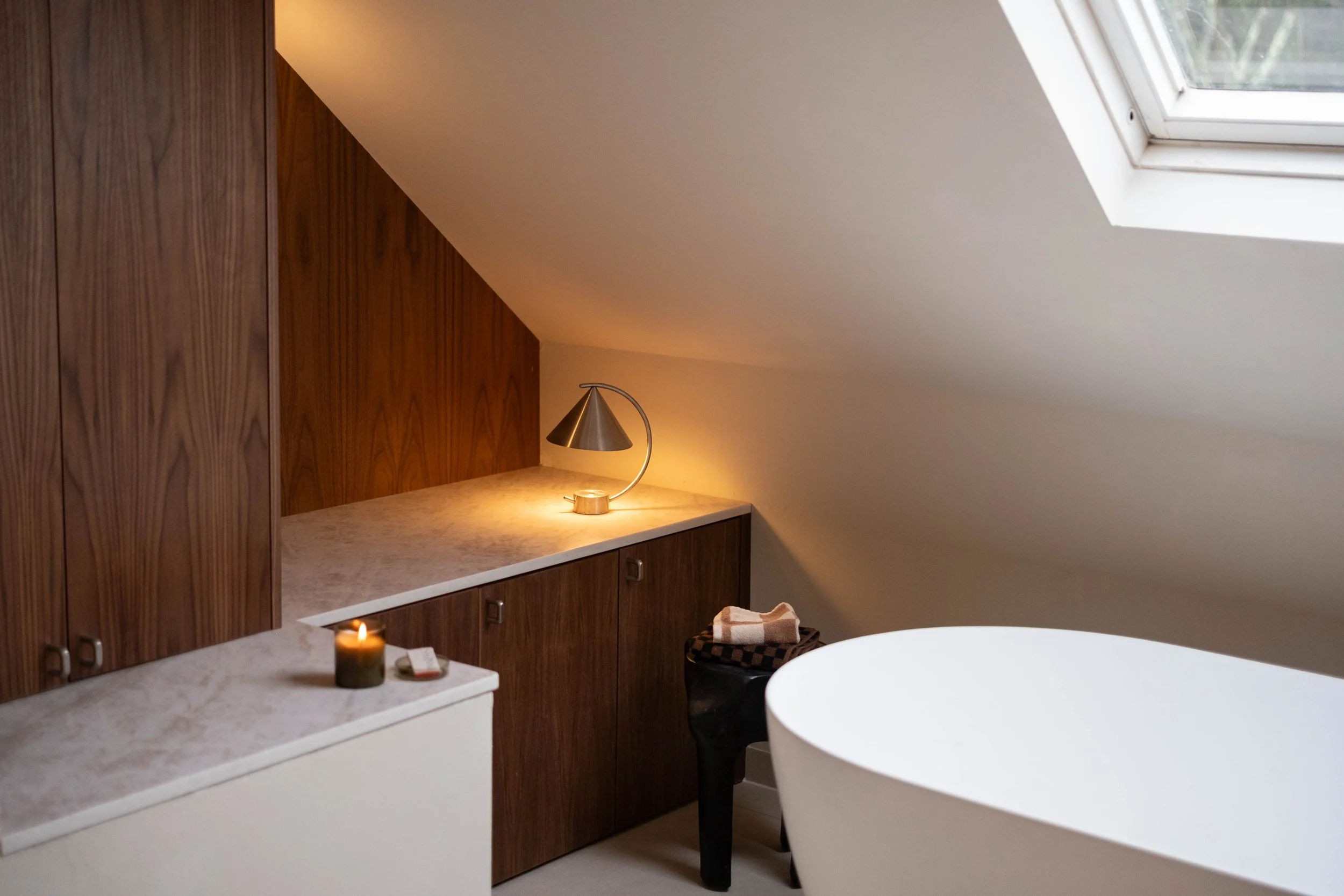 A cozy room with a sloped ceiling, featuring a wooden cabinet, a small table with a lit candle, a modern desk lamp, a basket with a blanket, and a white, oval-shaped bathtub under a skylight window.