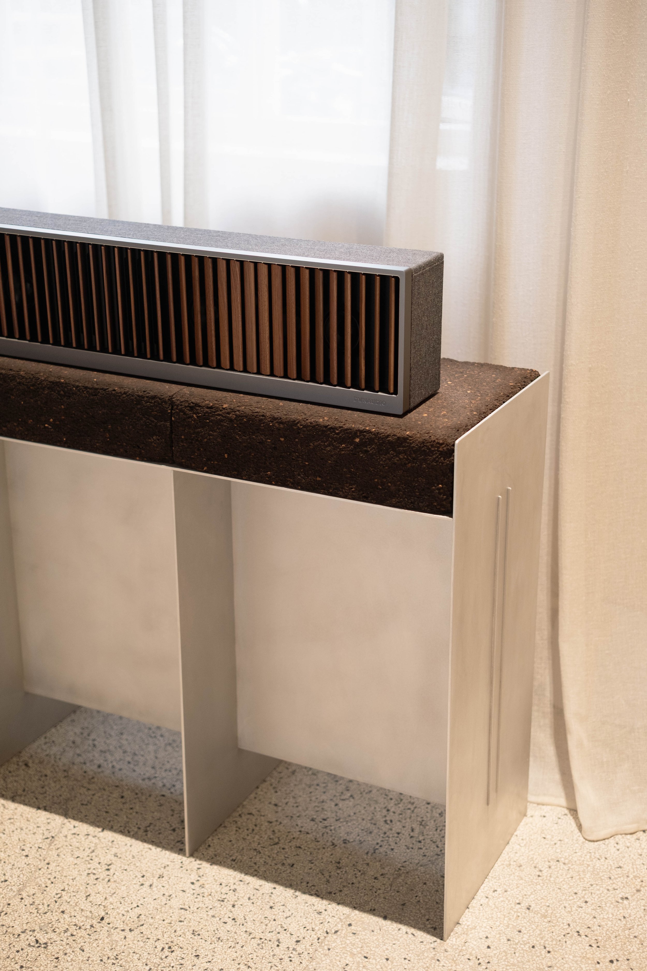 A speaker with a black grille and wooden slats, placed on a white cabinet. Behind it, sheer curtains are partially visible over a window.