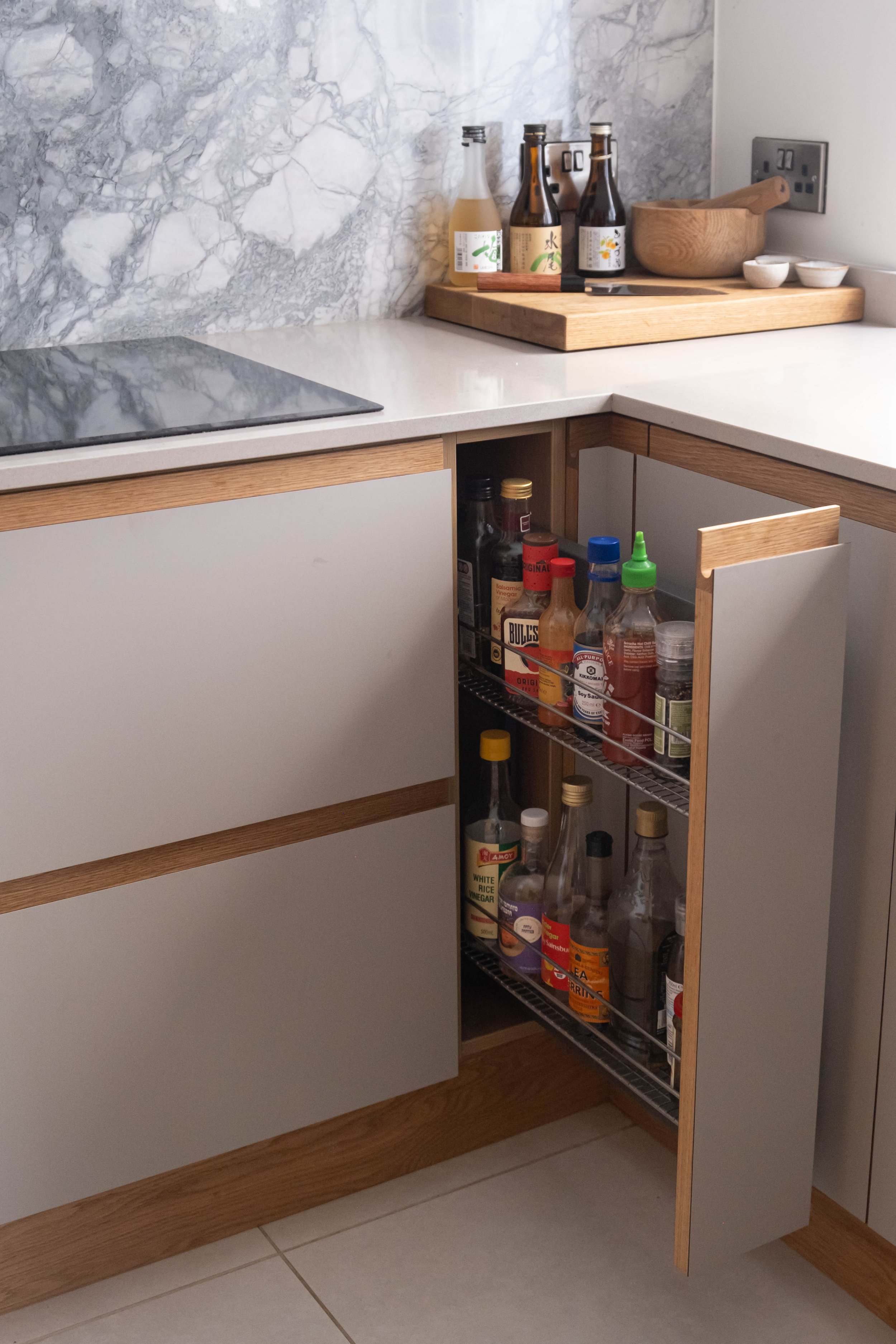 Kitchen corner with a wooden cupboard, white countertop, marble backsplash, open spice rack with bottles and jars, and a wooden bowl on a cutting board.