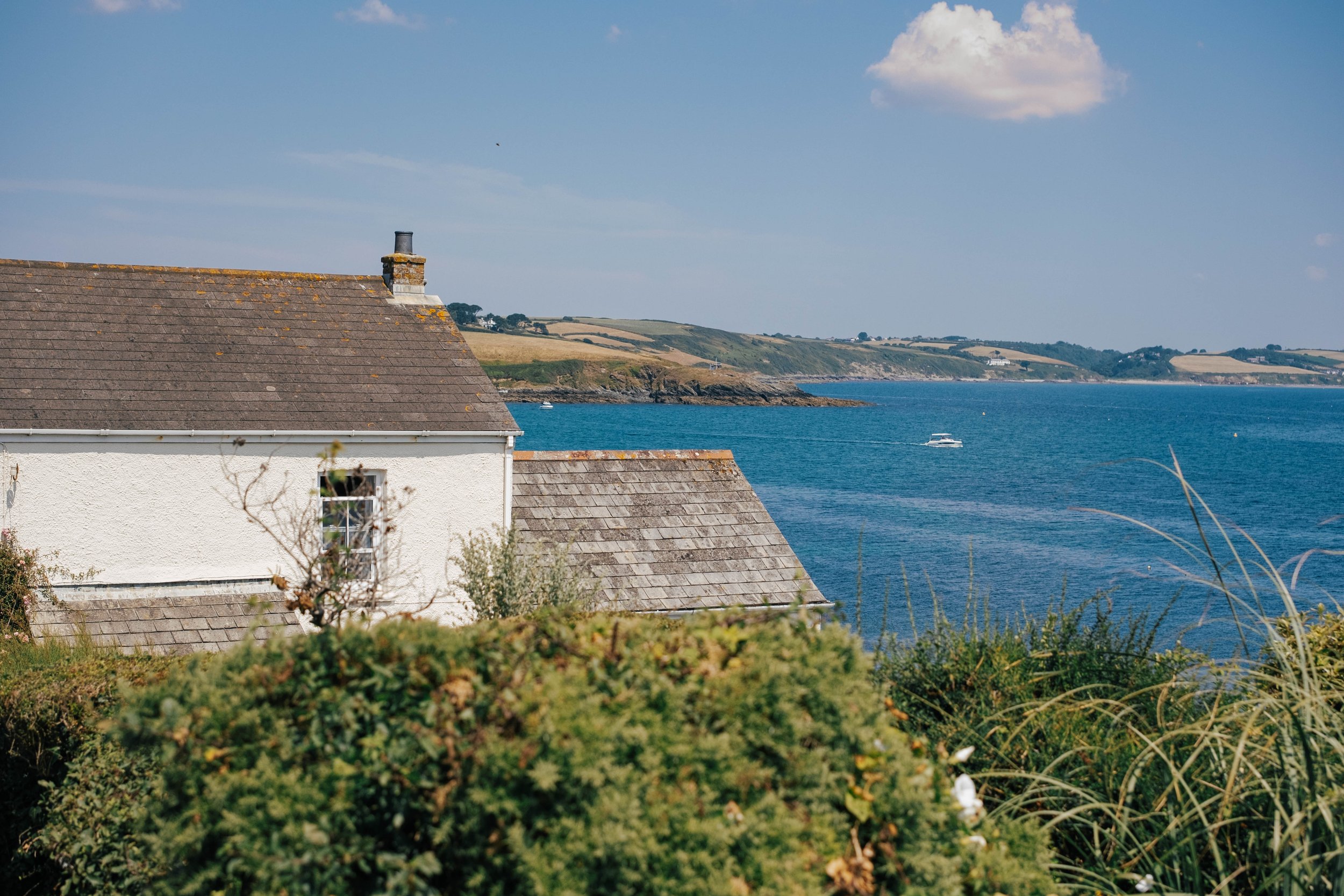 A house with a sloped shingle roof overlooking a body of water with boats, distant hills, and partly cloudy sky.