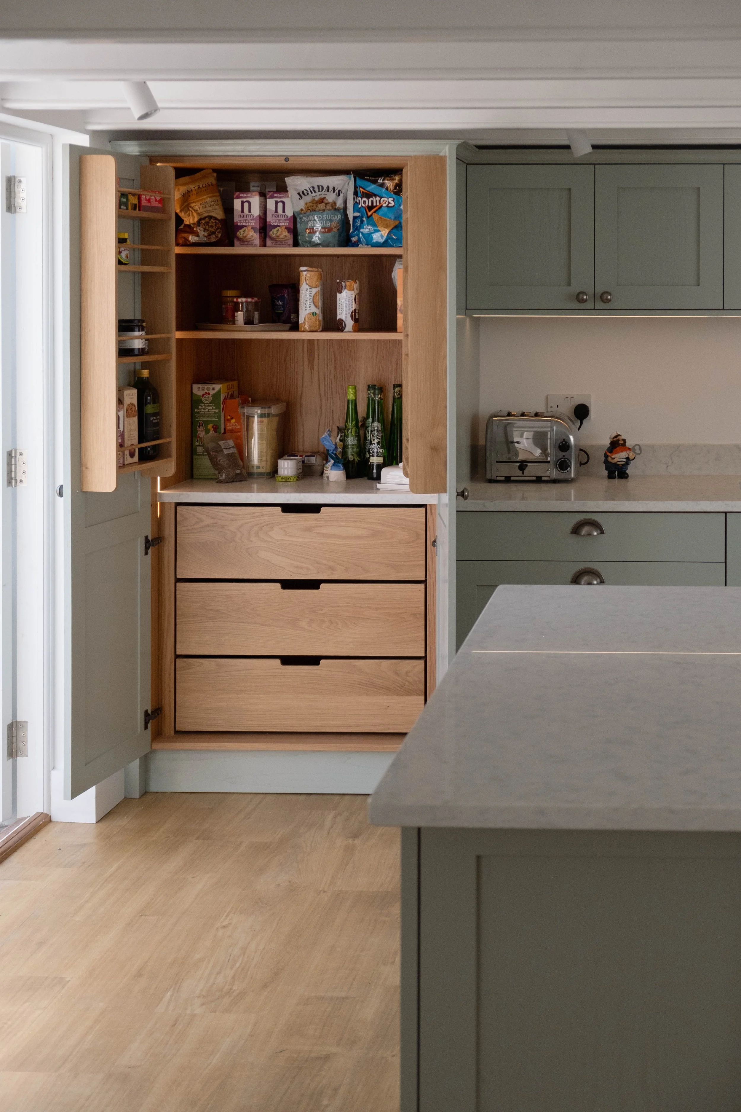 Open kitchen cabinet with wooden drawers and shelves stocked with snacks, jars, bottles, and containers, next to gray kitchen cabinets, countertop with toaster, and a small figurine.