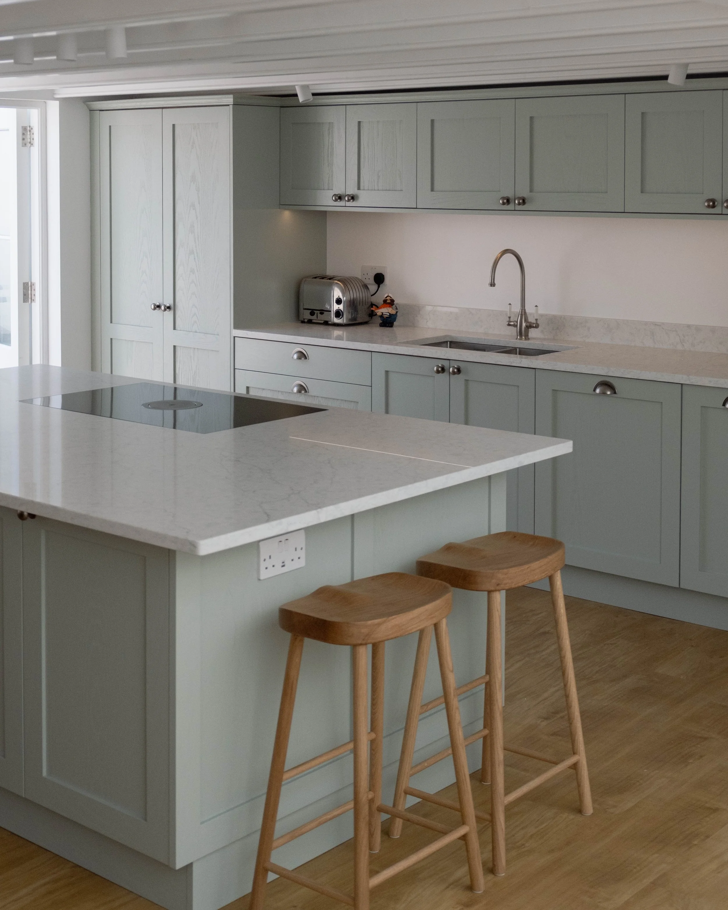 A modern kitchen with light grey cabinets, a white marble countertop, a built-in stovetop, a stainless steel toaster, a small decorative figurine, and wooden bar stools by the island. There's a window and a door to the outside in the background.
