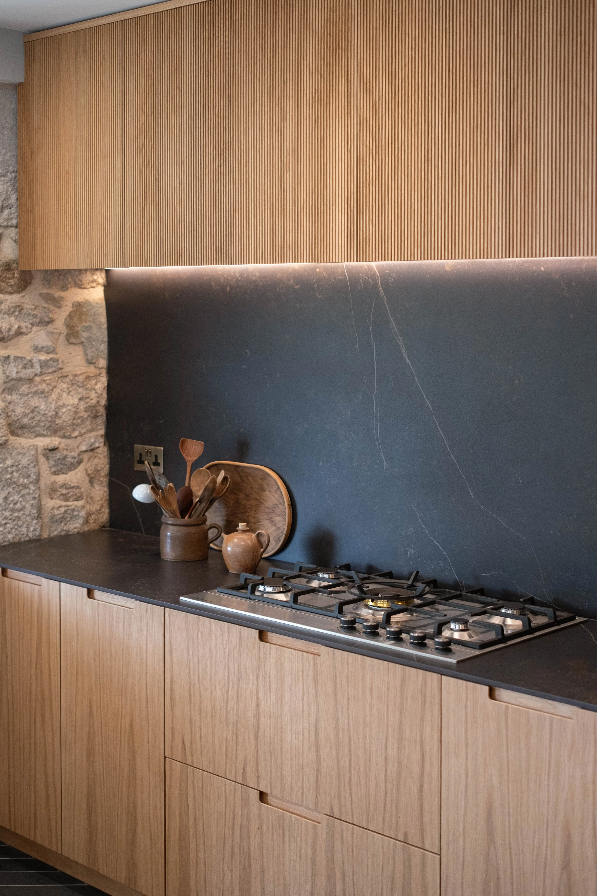 Kitchen with wooden cabinets, a black countertop, a black marble backsplash with white veins, a gas stove, and decorative items including a jug filled with wooden cooking utensils and two small ceramic pots.