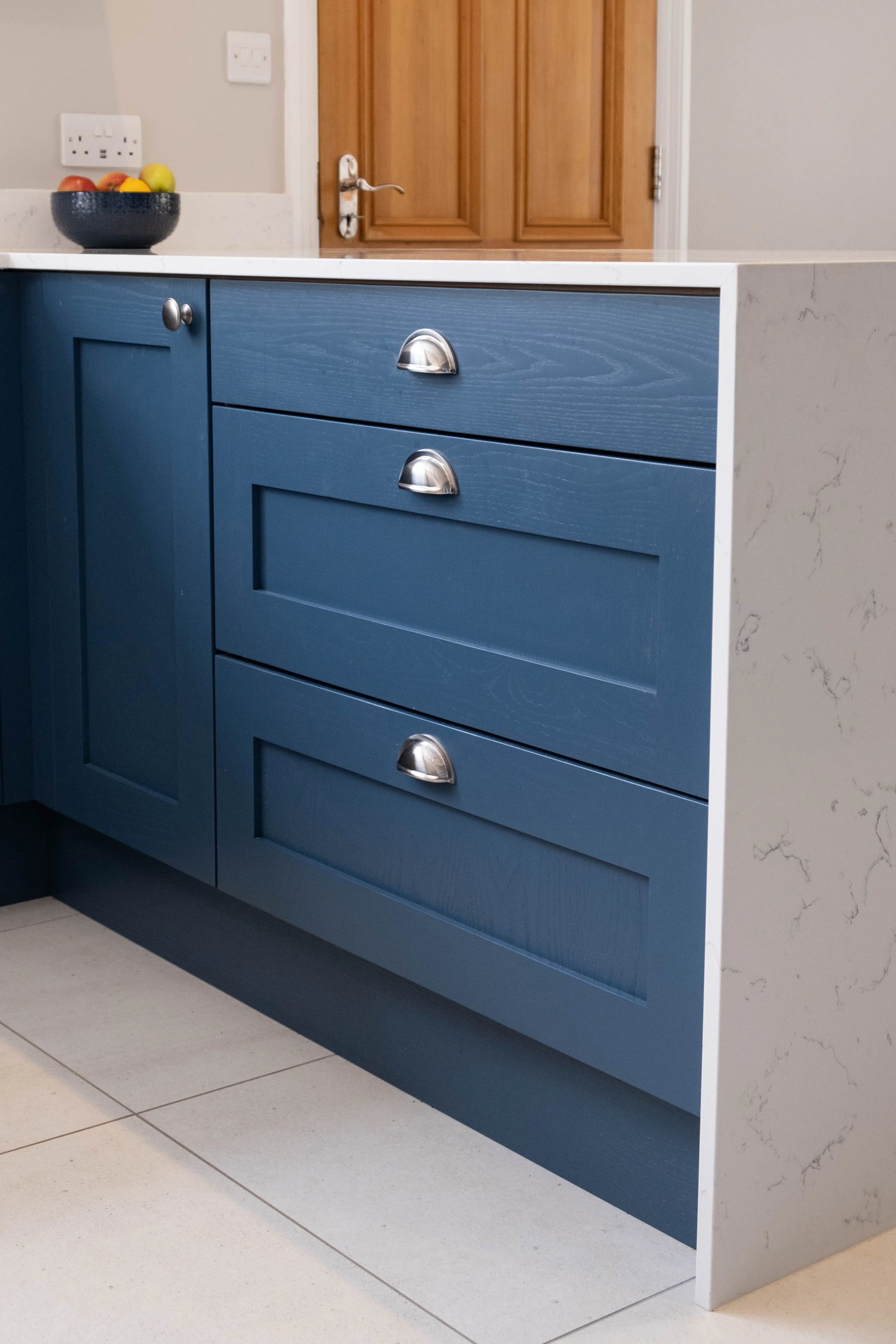Blue kitchen cabinet with silver handles and a marble countertop, with a bowl of fruit on top, against a wall with electrical outlets and a wooden door in the background.
