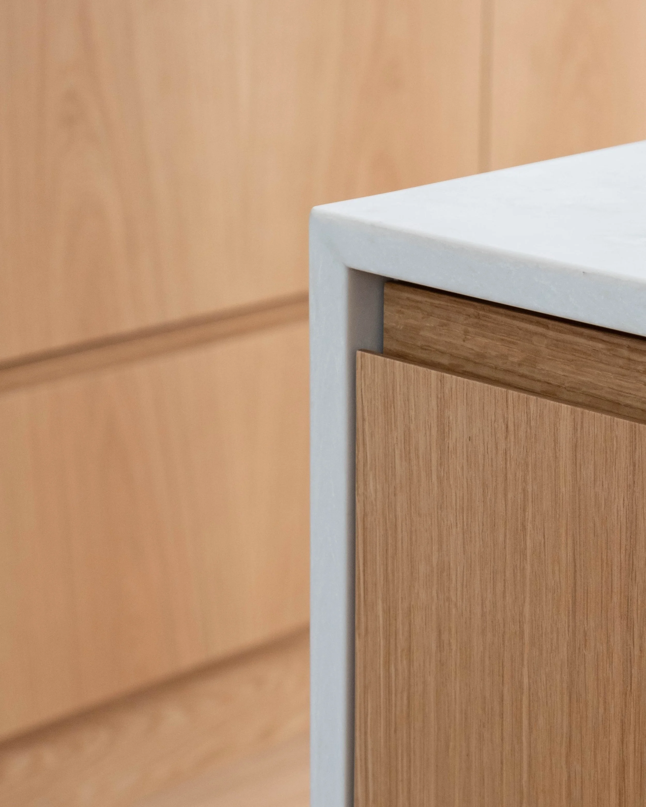 Close-up of a corner of a wooden furniture piece with a white top surface, set against a light brown wooden panel background.