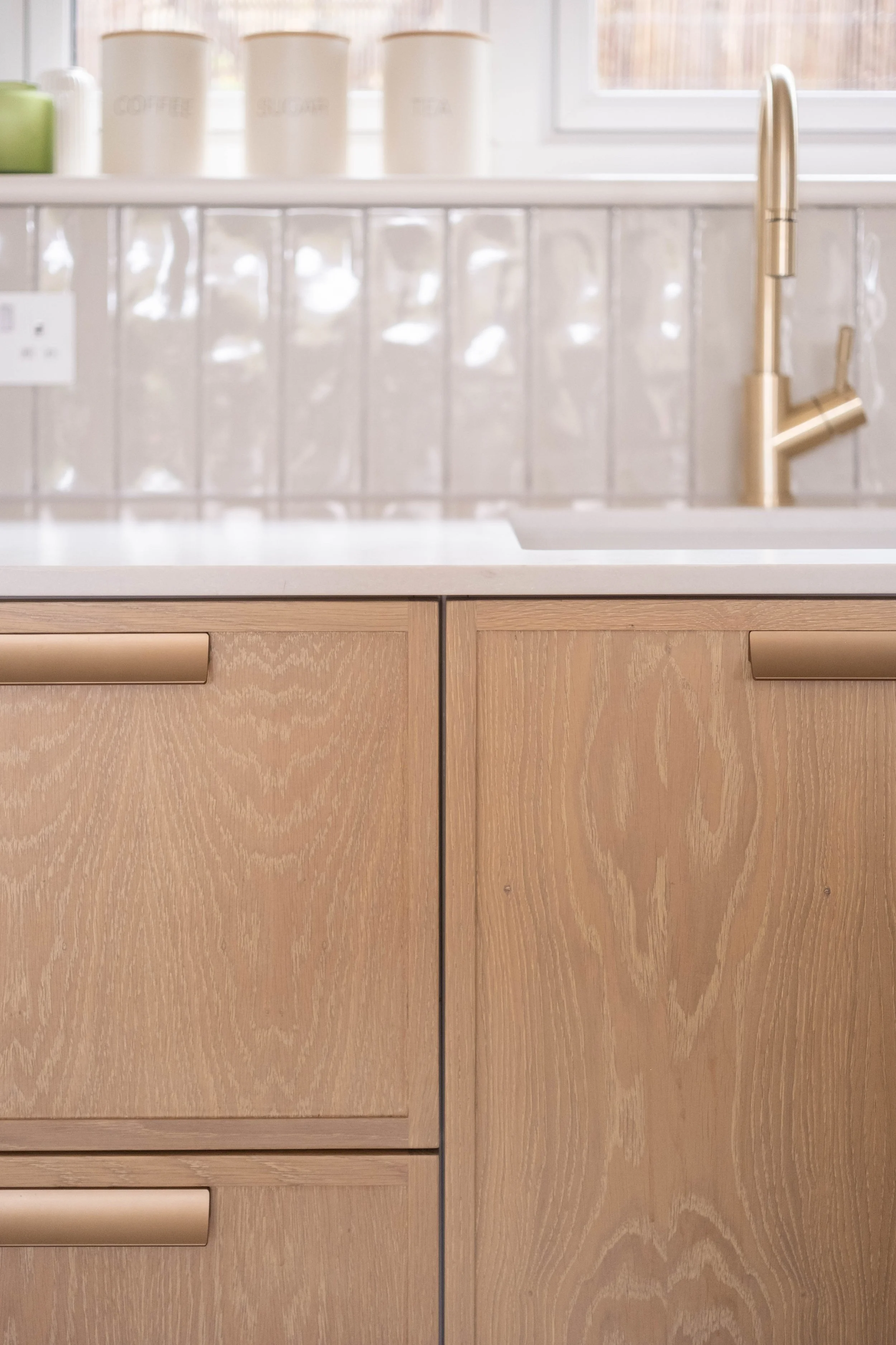 Kitchen with wooden cabinets, a white countertop, a gold faucet, and white jars labeled 'Coffee,' 'Sugar,' and 'Tea' near a window.