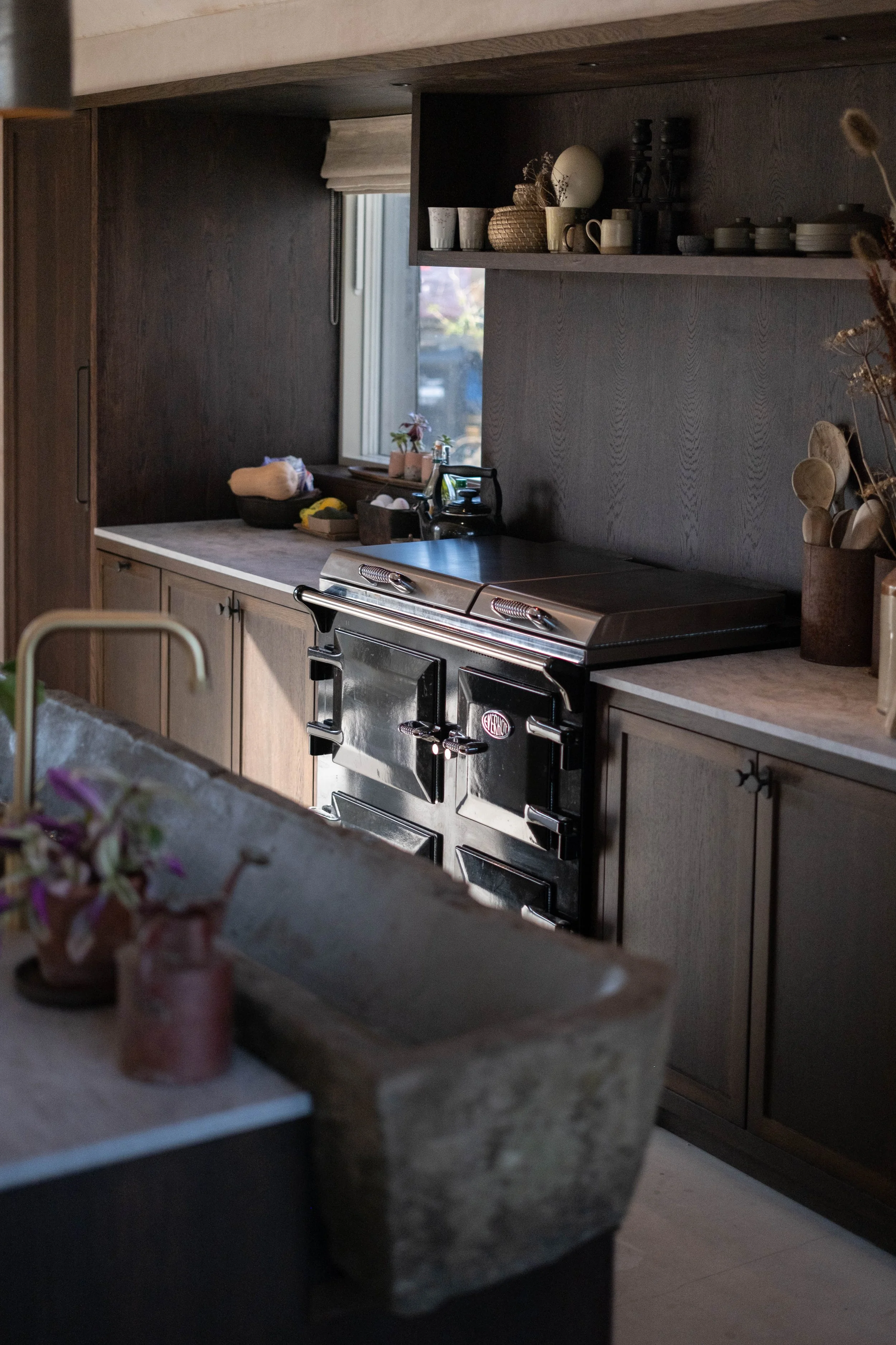 A kitchen with dark wood cabinets, a window with a roller blind, a stovetop, and a countertop with a small potted plant, kitchen utensils, and dishes on the shelves.