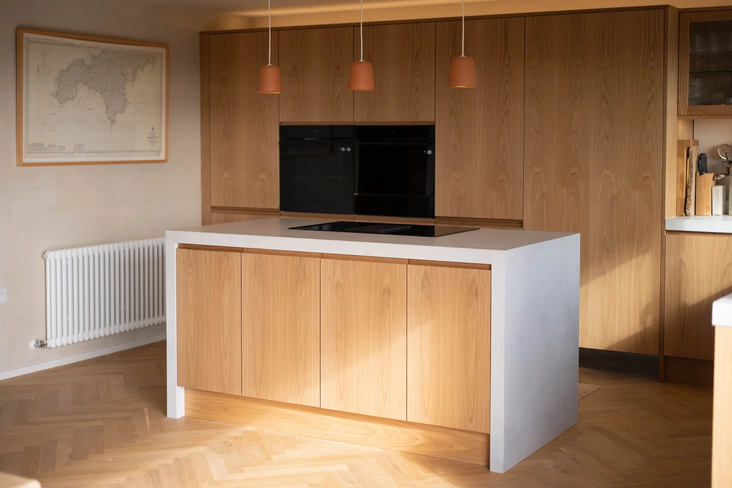 Modern kitchen with wooden cabinets and white island counter. Hanging pendant lights above the island. Map of Spain on the wall, radiator on the wall, and some kitchen utensils on a small counter.