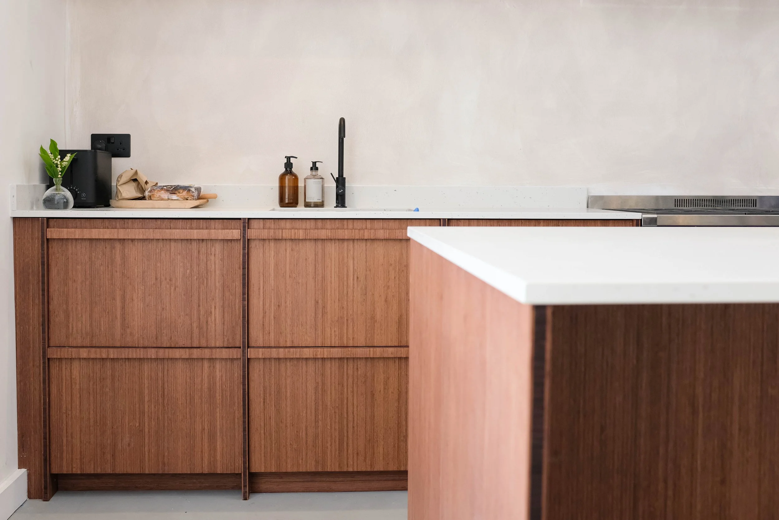 Kitchen with wooden cabinets, white countertop, black faucet, and small decorative items including a glass vase with green leaves and white flowers, brown soap dispenser, and a small brown bottle.