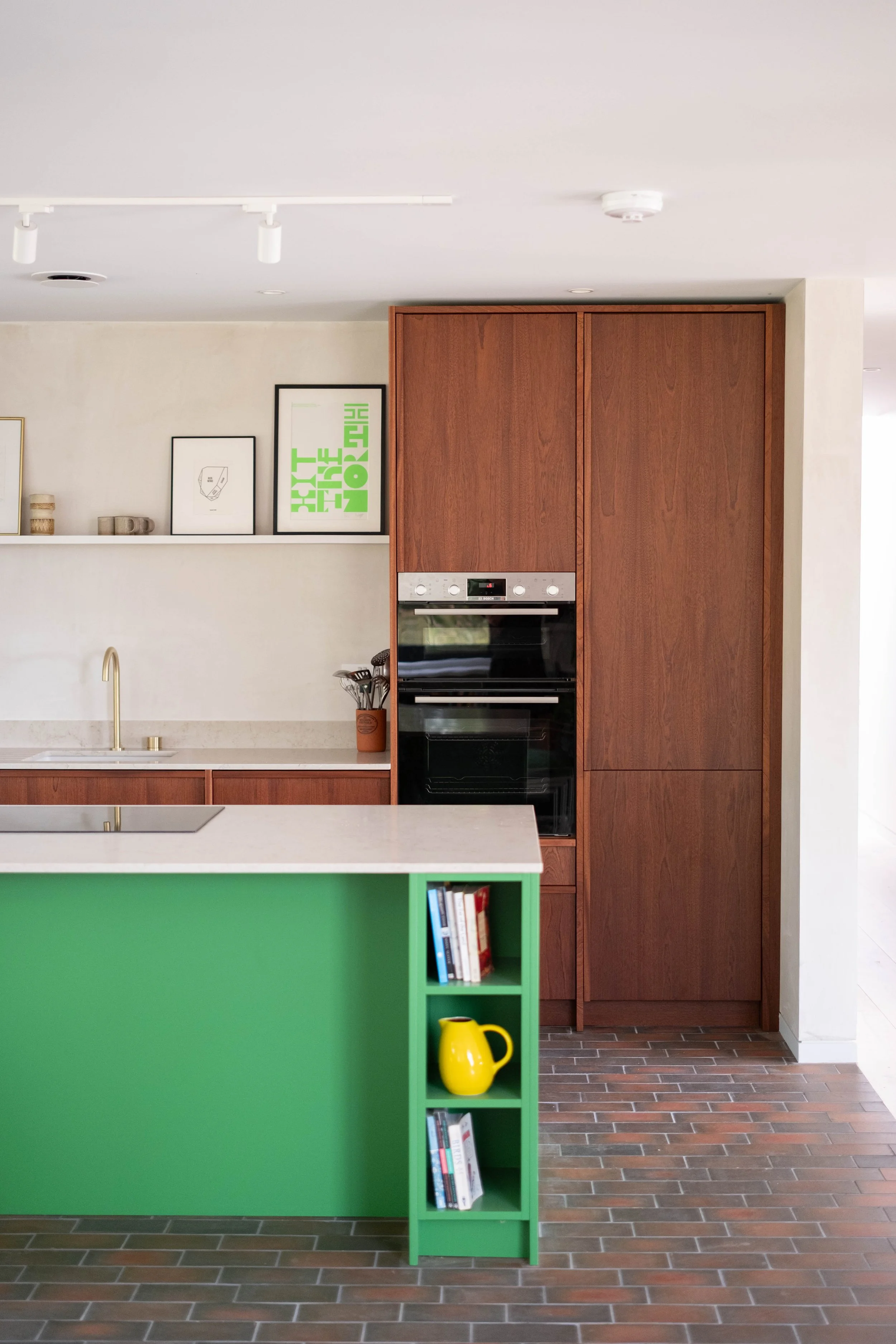 A modern kitchen with a white countertop, a wooden cupboard, and a green bookshelf with books and a yellow pitcher. The wall has framed artwork, and the floor has wood-like tiles.