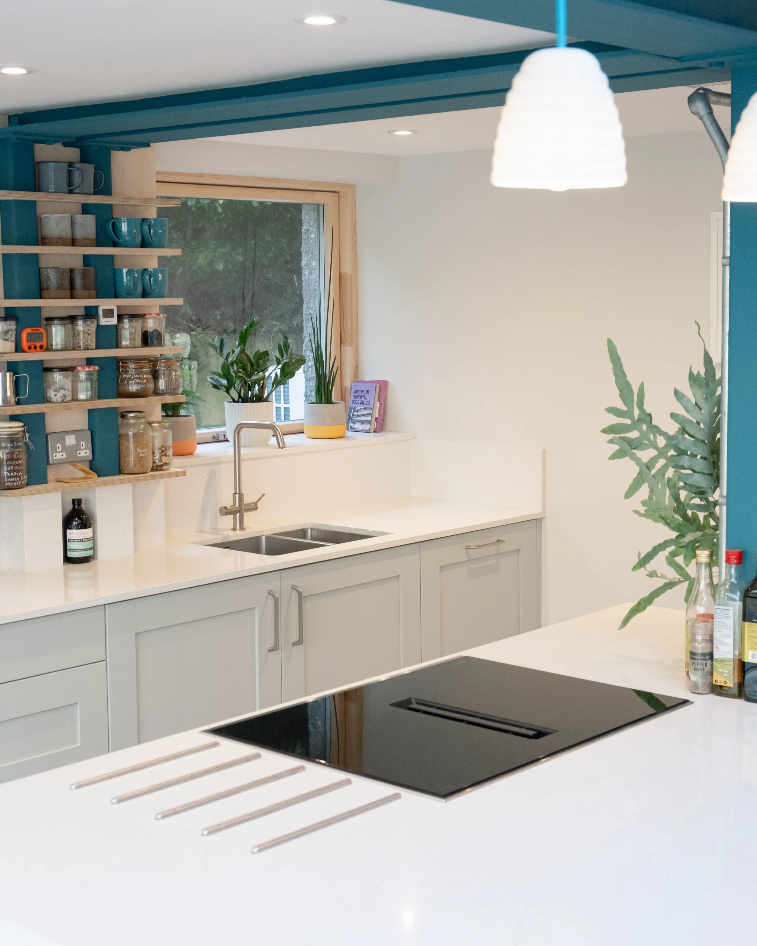 Modern kitchen with white countertops, light blue cabinets, a black stovetop, potted plants, open shelving with cups and jars, and a large window with greenery outside.