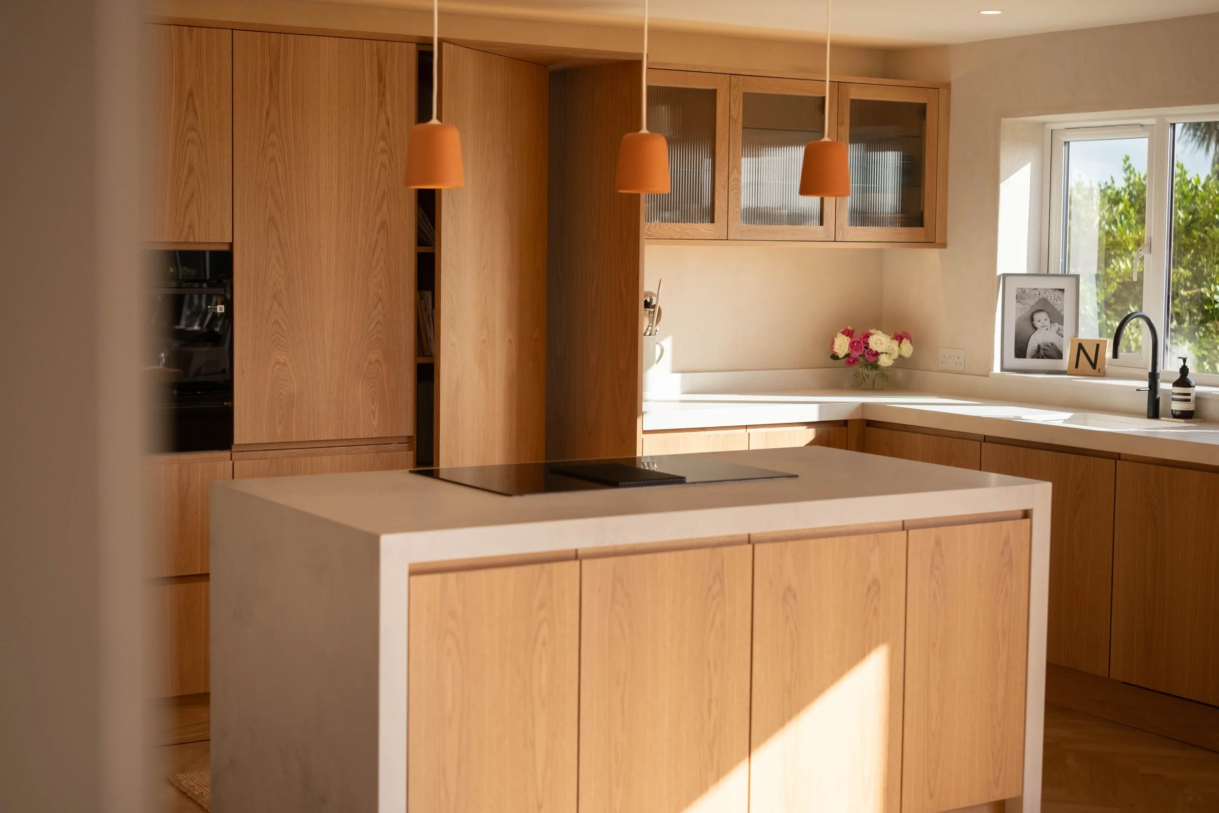 A modern kitchen with wooden cabinets, a white kitchen island, and a window with greenery outside. Decor includes a framed black and white photo, a vase of pink and white flowers, a letter 'N', and a soap dispenser near the sink.