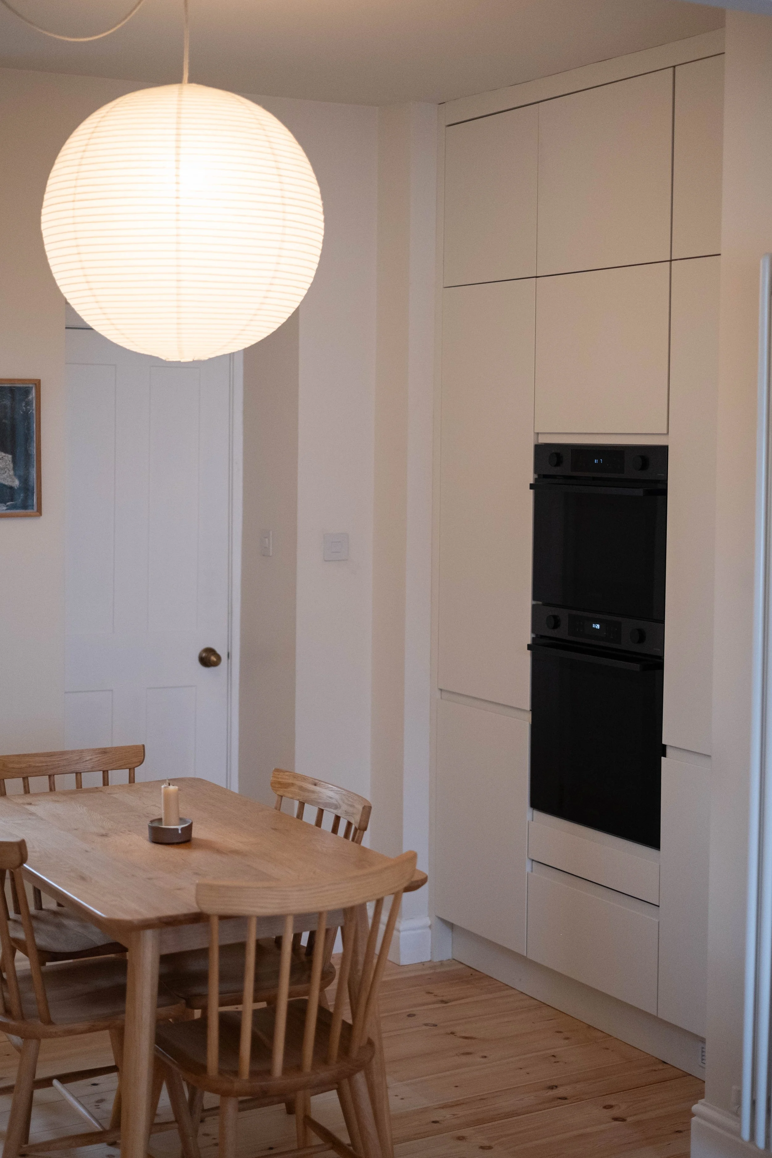 A dining room with a wooden table and chairs beneath a large round paper lantern hanging from the ceiling. In the background, there is a white cabinet with built-in black ovens, and a wall with a white door and artwork.