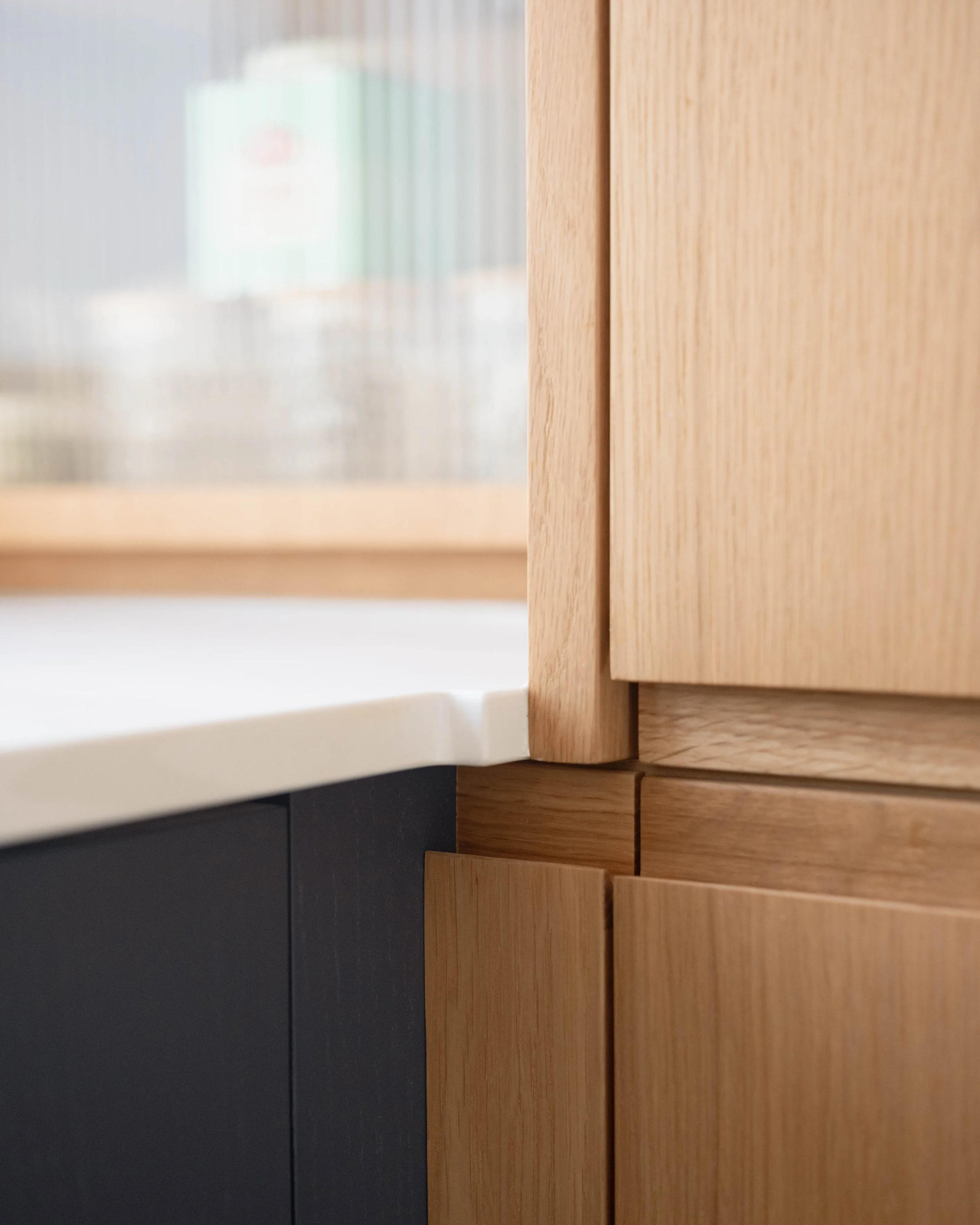 Close-up view of a wooden kitchen cabinet with a white countertop, a light-colored wooden door, and a dark gray cabinet below, with a blurred background.
